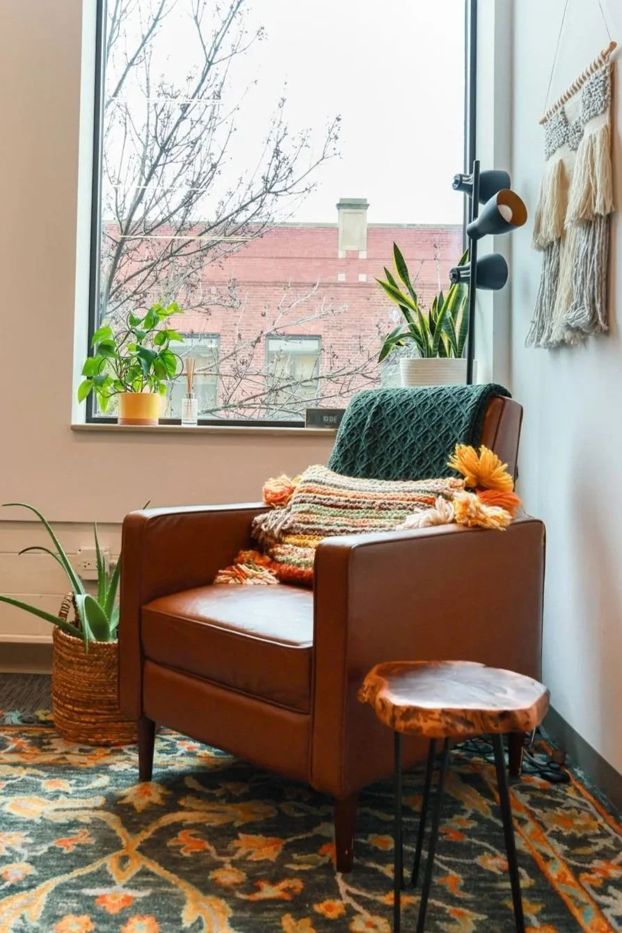 A cozy corner in a room with a brown leather armchair draped with colorful throws and pillows, a small wooden side table, a large window with potted plants outside, and a decorative wall hanging, rug, and floor lamp inside.