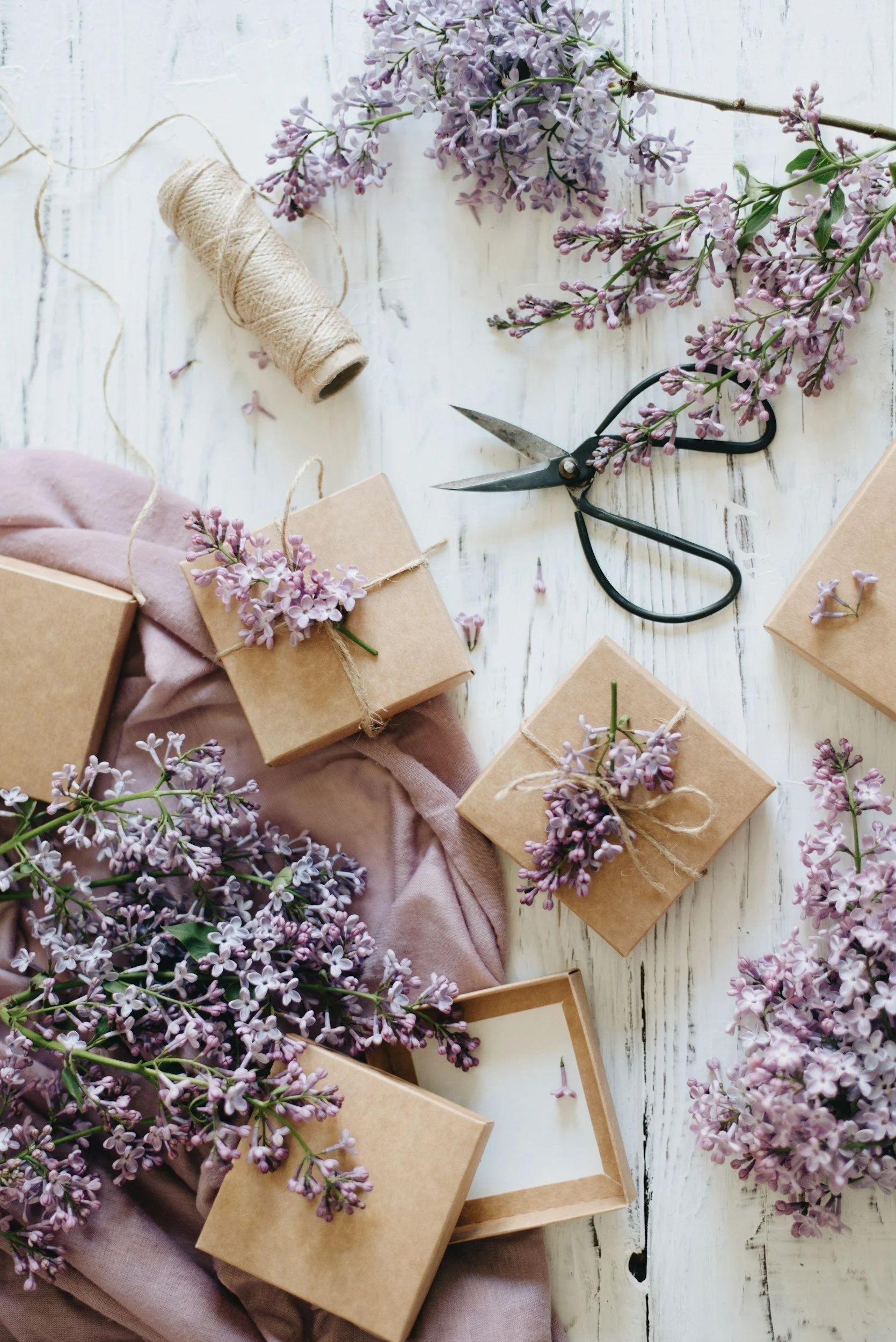 Flat lay of small gift boxes wrapped in brown paper with purple lilac flowers, a pair of scissors, and natural twine on a white wooden surface with lilac flowers scattered around.