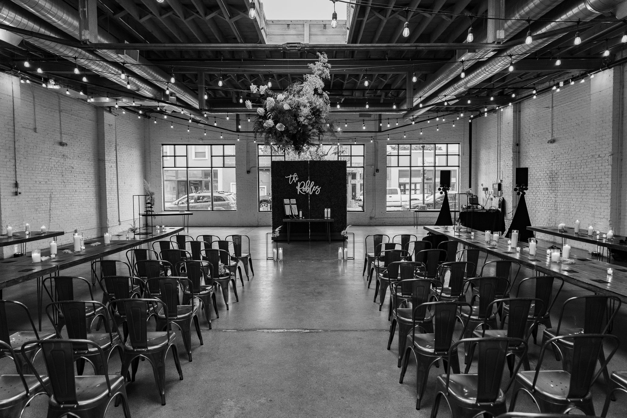 An indoor event space decorated for a celebration, with chairs and tables arranged around a dance floor, string lights hanging from the ceiling, a floral arrangement hanging above, and candles on the tables, with large windows at the back showing par