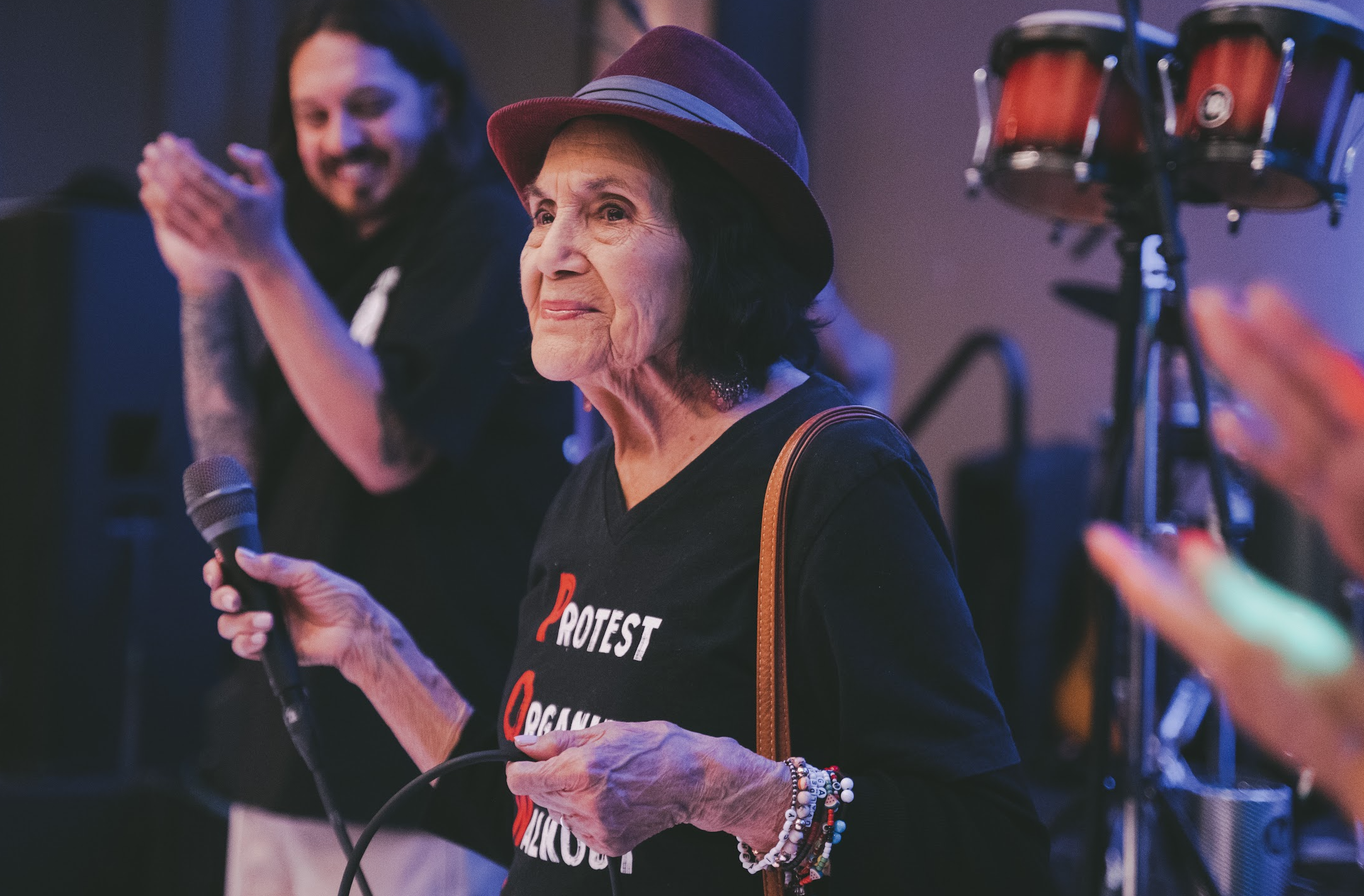 An elderly woman holding a microphone at a protest event, wearing a black shirt with the words 'Protest' and 'Reclaim' visible, and a purple hat. She has bracelets on her wrist and is standing next to a drum set, with another woman clapping in the ba