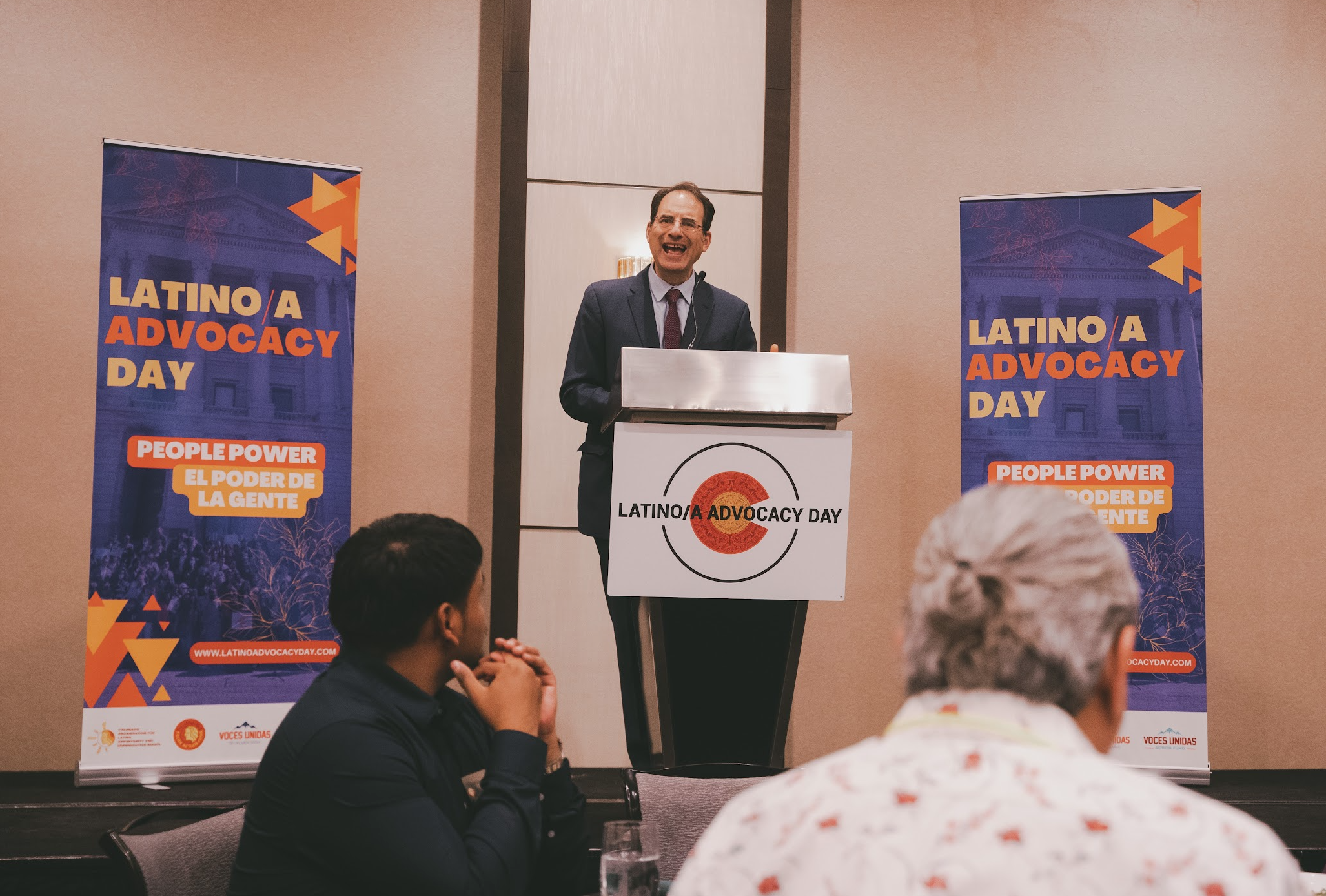 Man speaking at a podium during Latino/a Advocacy Day event, with two banners on either side reading "Latino/a Advocacy Day" and "People Power El Poder de La Gente." People seated in the audience watching the speaker.