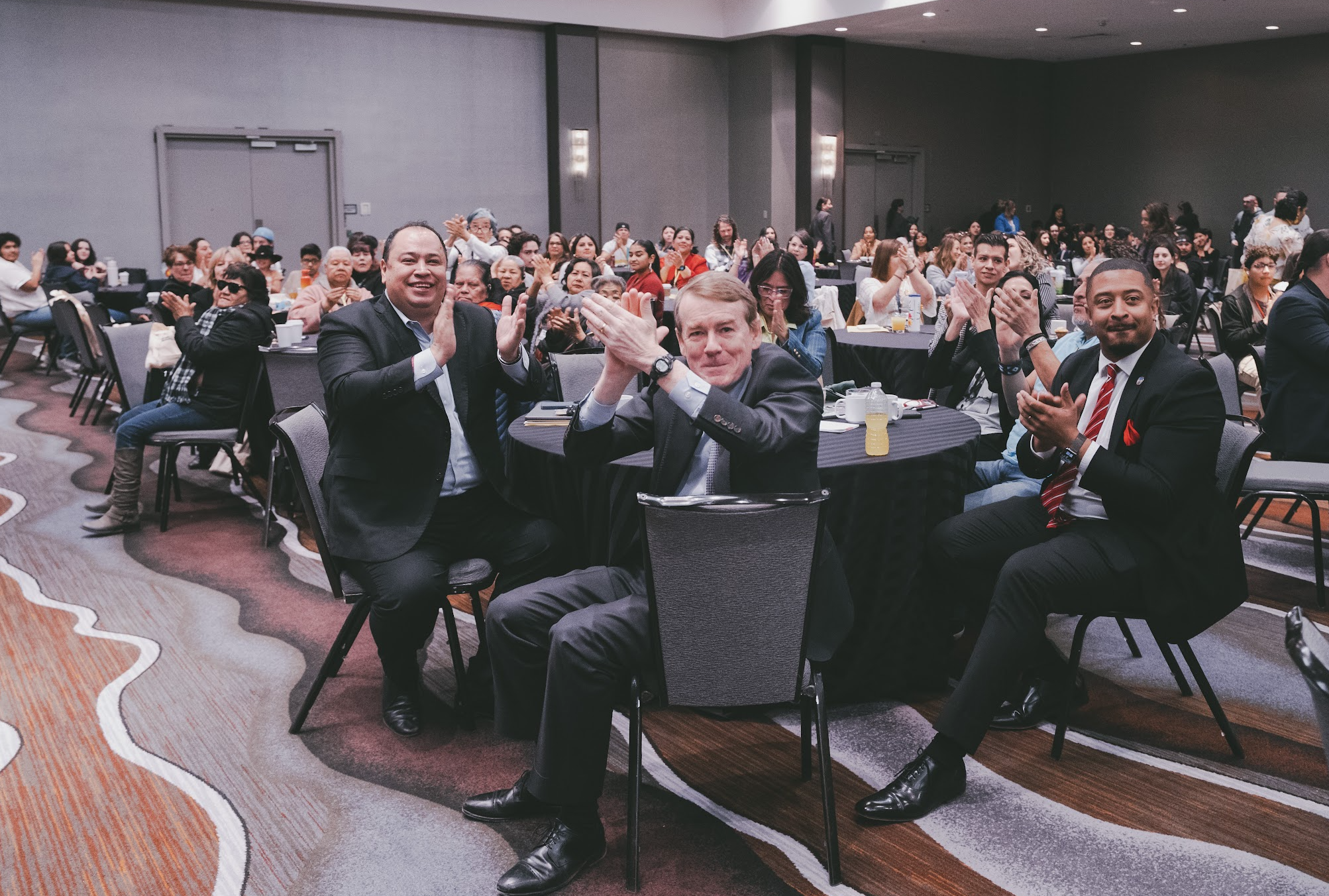 People at a formal event clapping and celebrating, seated around round tables in a large conference room.