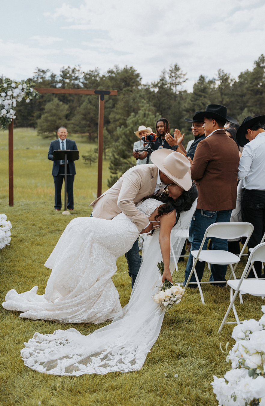 A couple sharing a kiss, the groom bending down while wearing a beige suit and hat, and the bride holding a bouquet, wearing a white lace wedding dress, during their outdoor wedding ceremony with guests in attendance, a officiant, and a photographer 