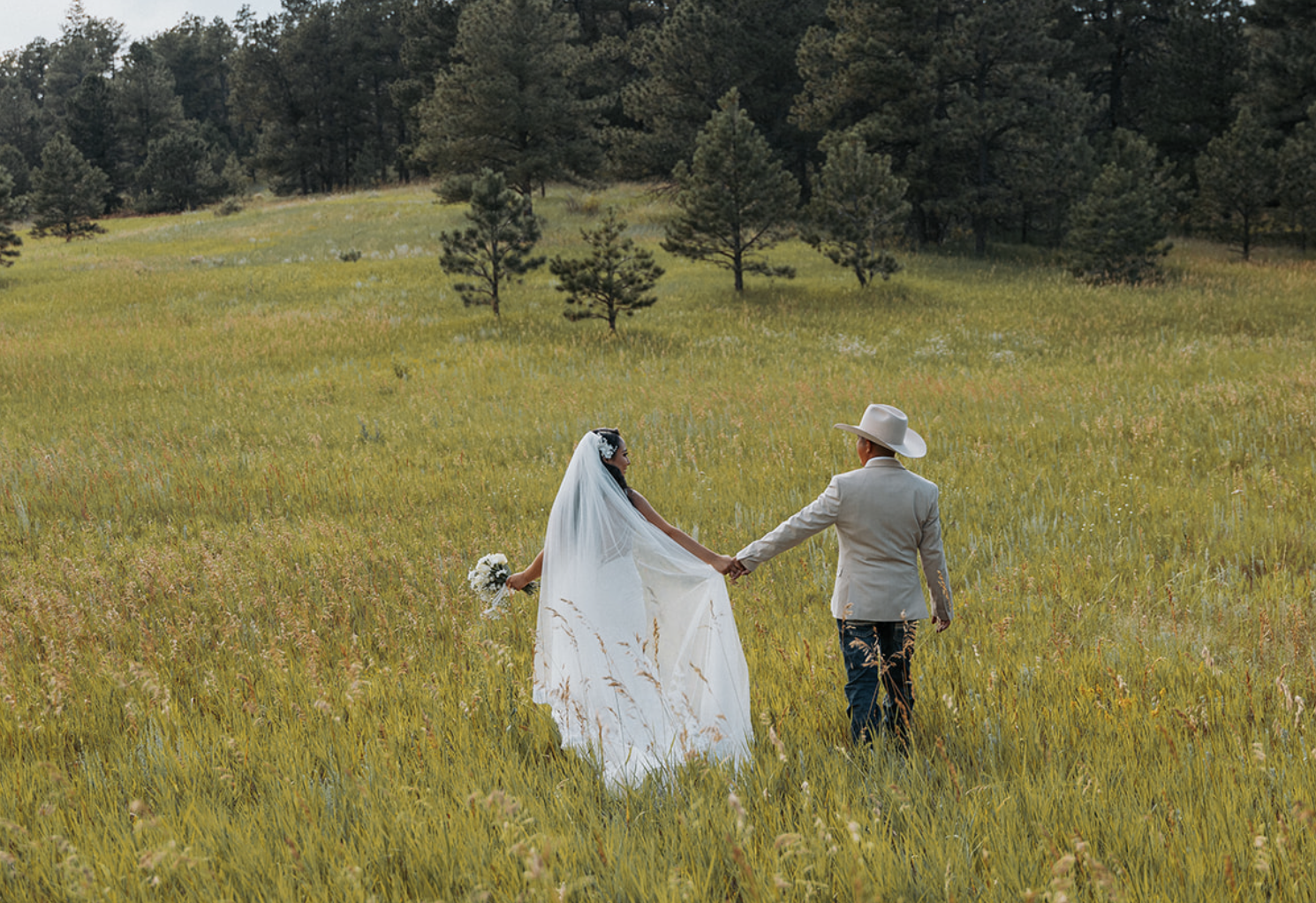 Bride and groom holding hands in a grassy field, with trees in the background.