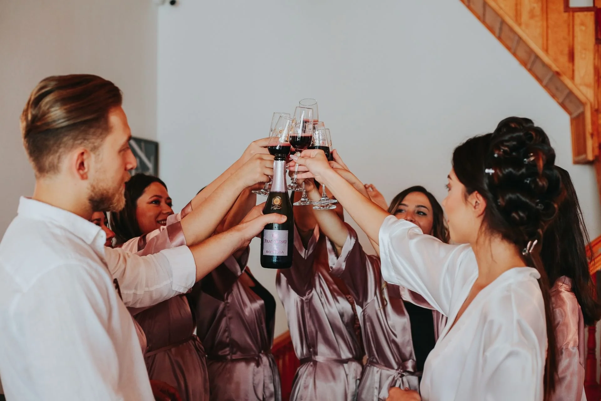 Group of women and men in pajamas raising glasses of red wine in a toast at a celebration or gathering in a cozy room with wooden ceiling panels.