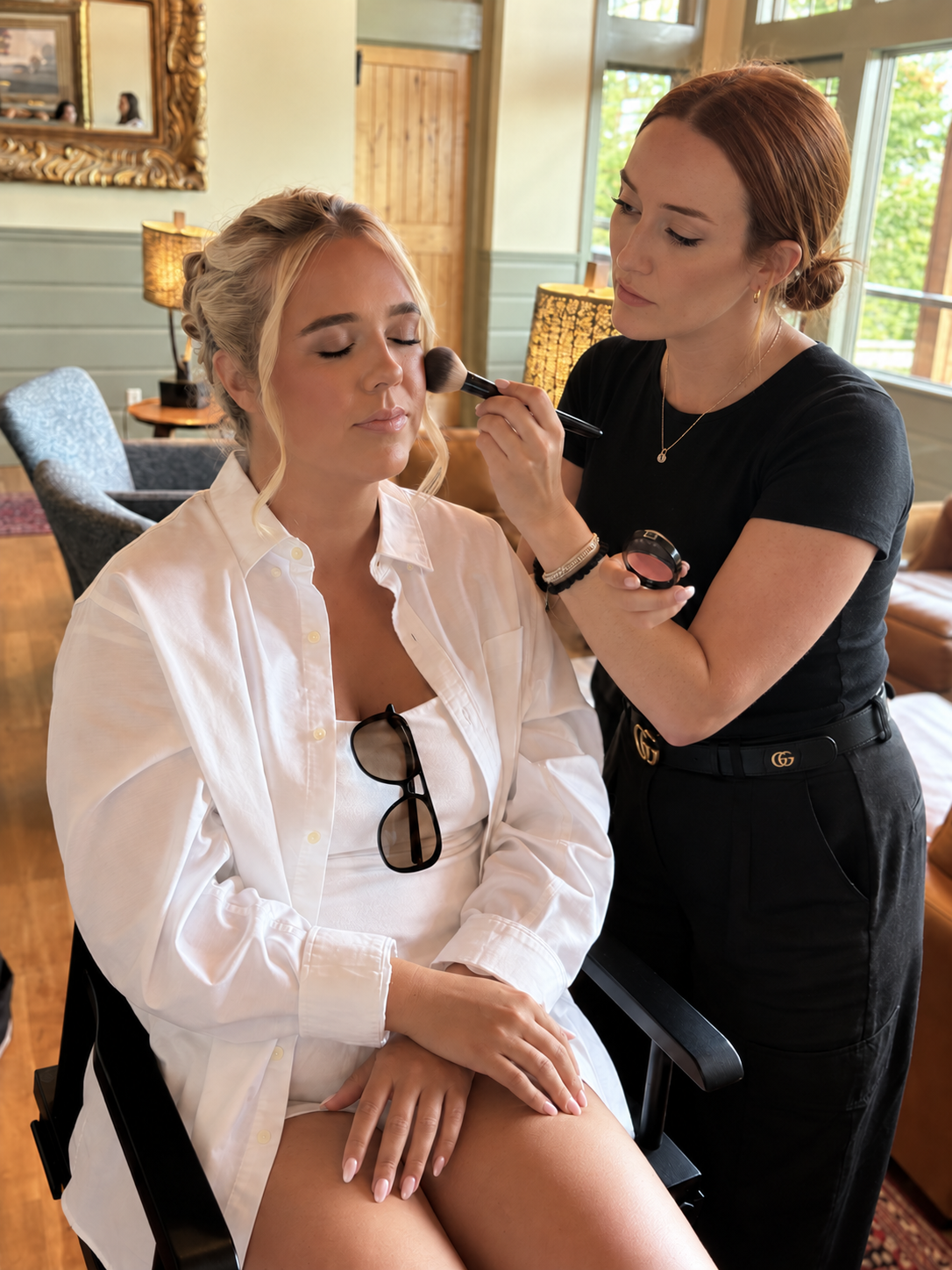 A woman sitting with eyes closed, receiving makeup application from a makeup artist in a cozy, well-lit room with wooden furniture and large windows.