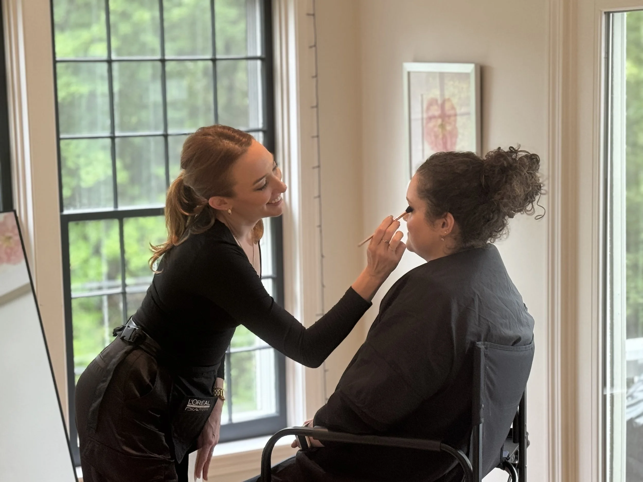 A makeup artist applying makeup to a woman sitting in a wheelchair in a room with large windows and framed artwork on the wall.