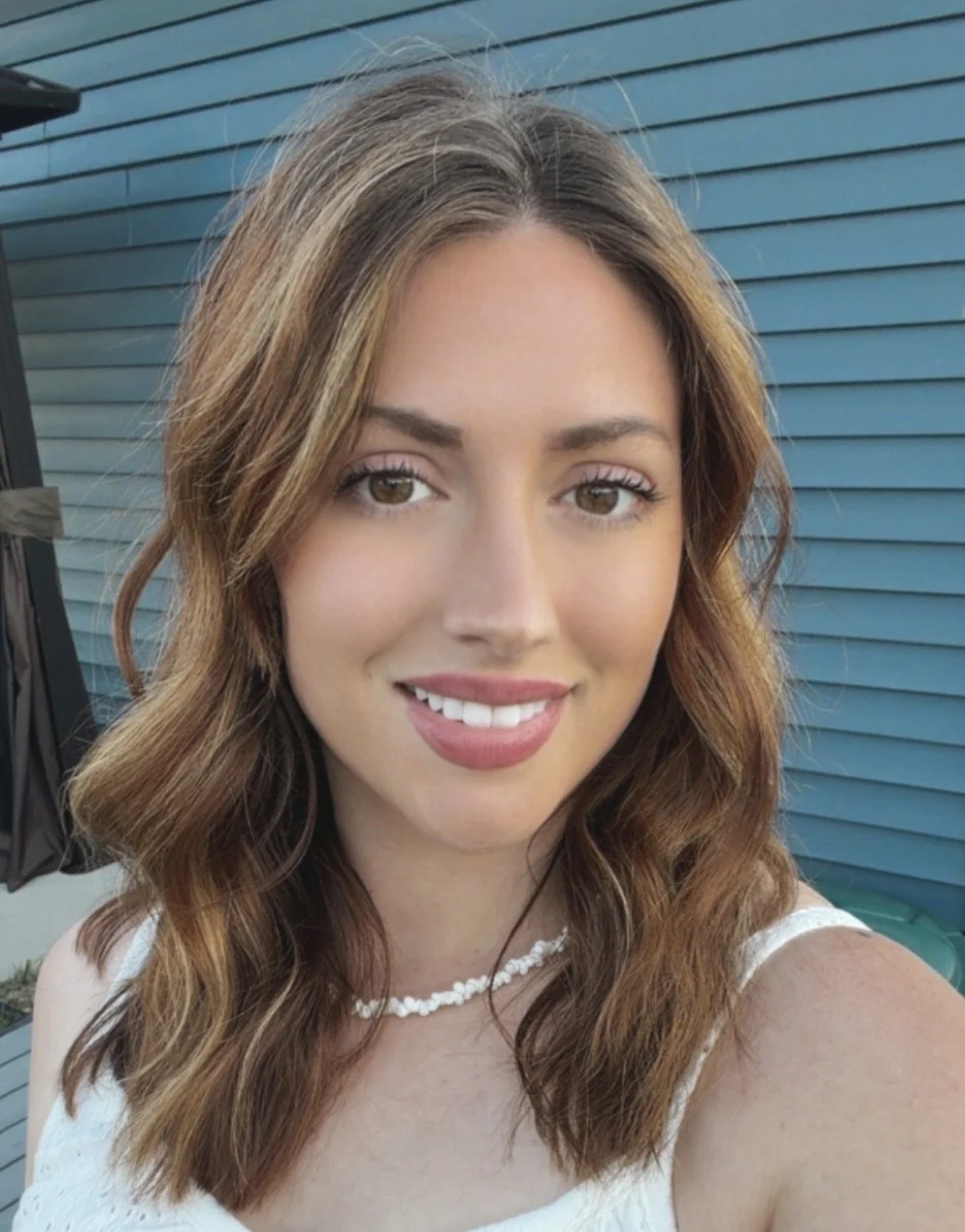 A woman with wavy brown hair, wearing a white top and a beaded necklace, smiling in front of a blue wall with horizontal siding.