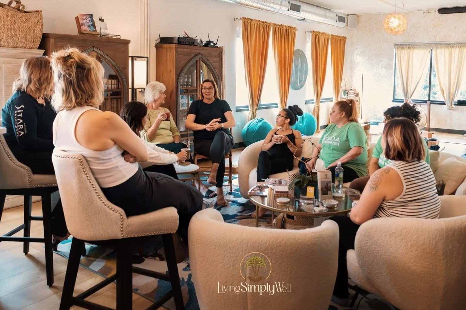 A diverse group of women gathered in a cozy, well-lit room for a discussion or workshop, seated on chairs and sofas, with drinks and notebooks on the tables.