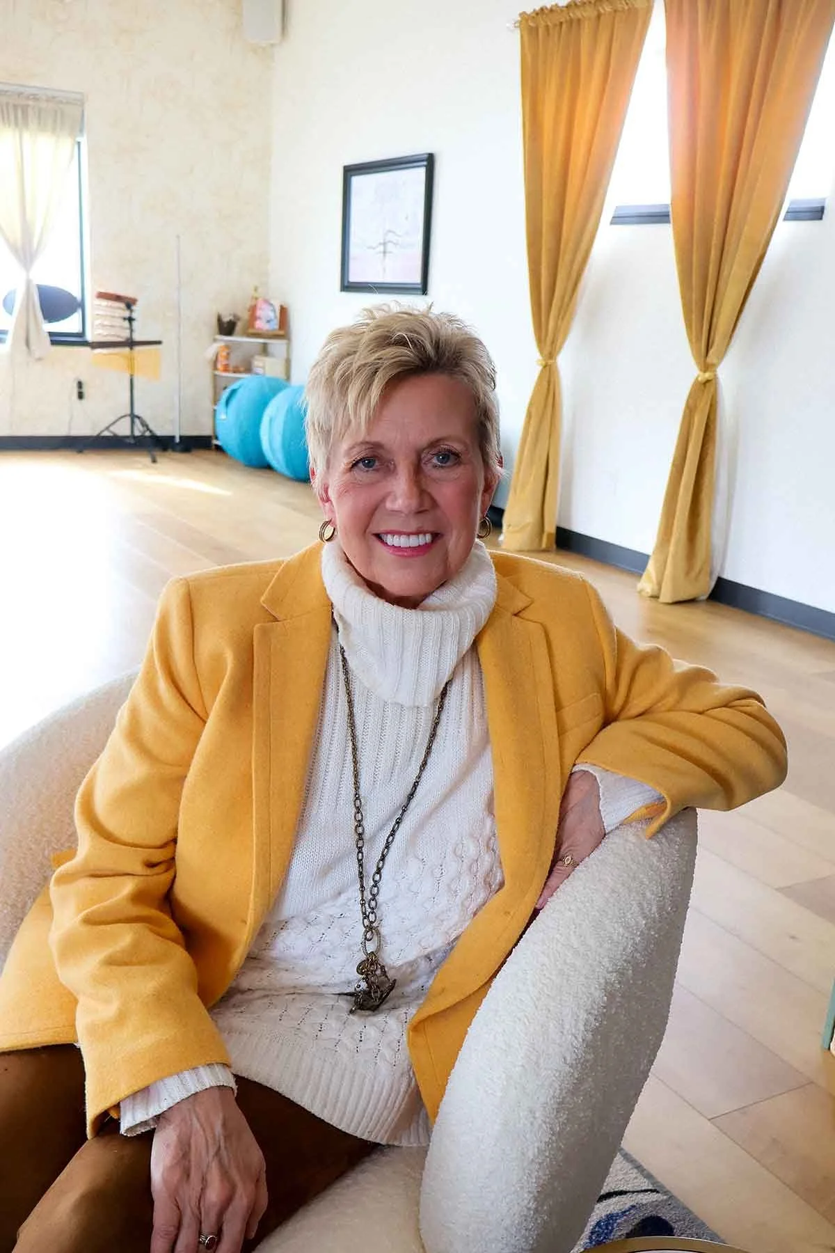 A smiling older woman with short blonde hair seated in a cream-colored armchair in a room with yellow curtains and light-colored walls. She is wearing a yellow blazer, cream turtleneck sweater, and a long necklace.