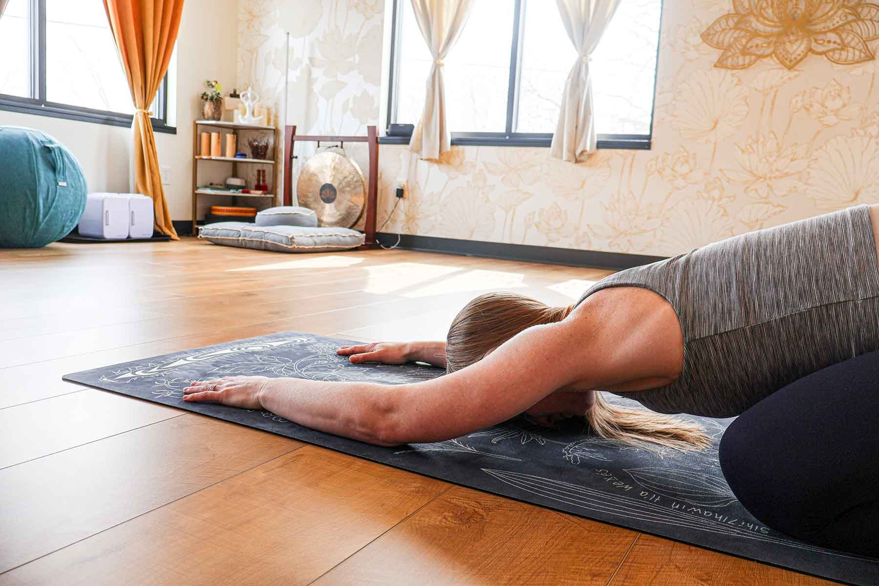 A woman practicing yoga in a room with wooden floors, stretching forward on a yoga mat with her arms extended and head down, in a child's pose.
