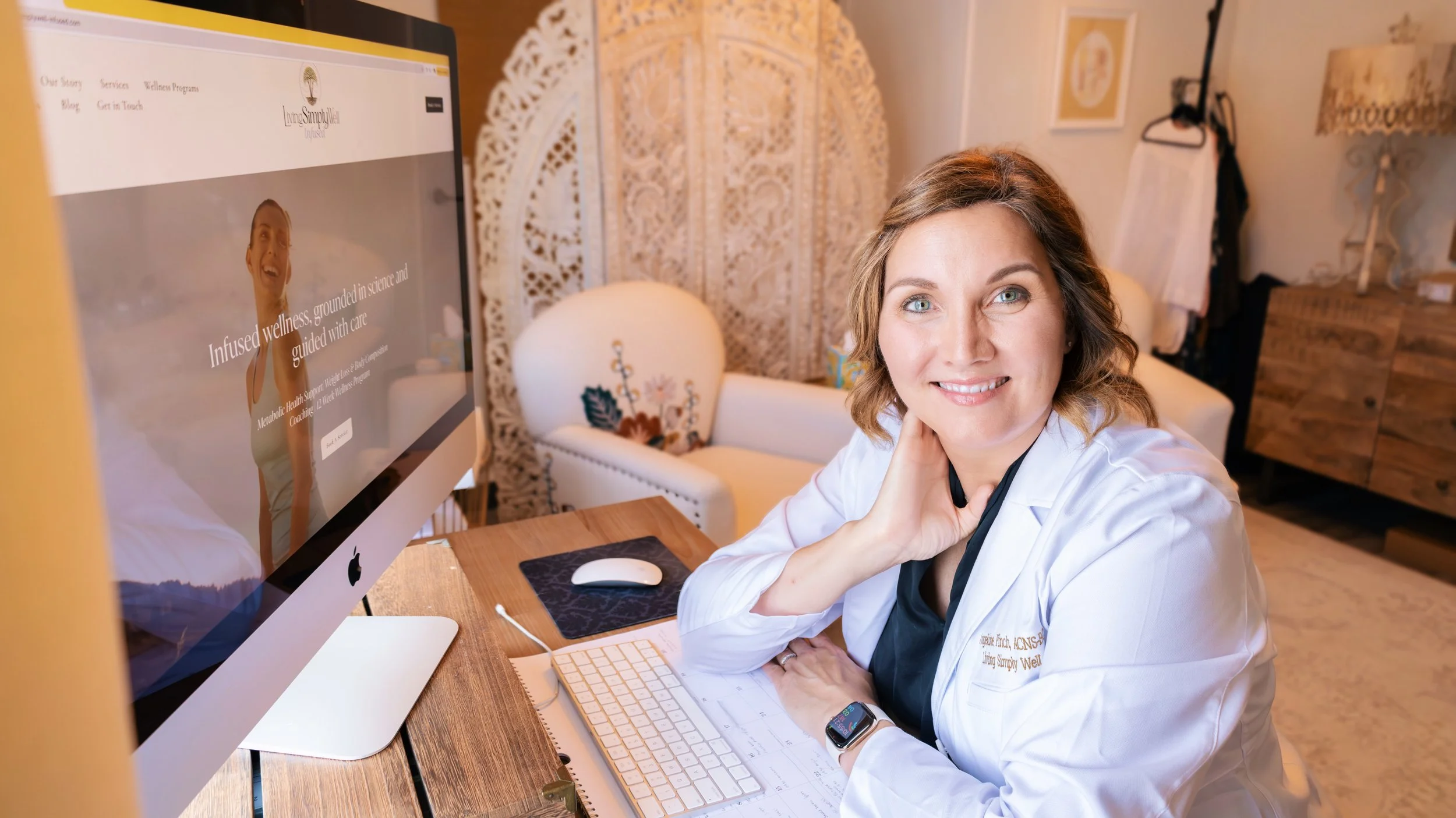 A woman in a white coat sitting at a desk in front of a computer, smiling at the camera. The computer screen displays a website related to wellness and science.
