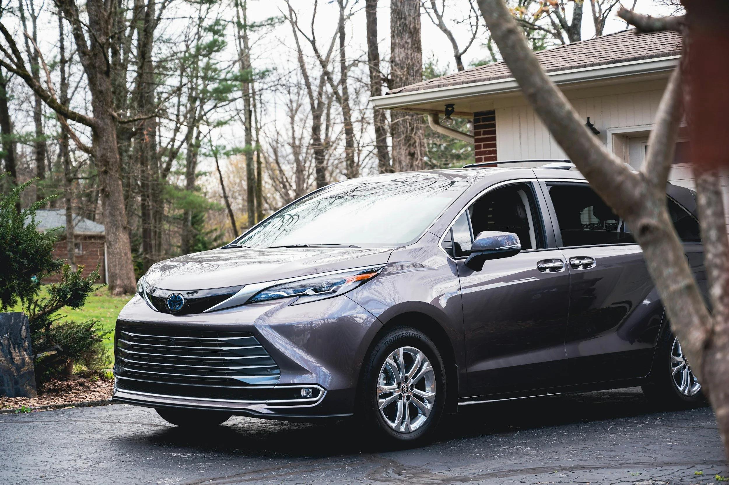 Gray Toyota minivan parked in a driveway near a house with tan siding and brick wall, surrounded by trees in a residential neighborhood.