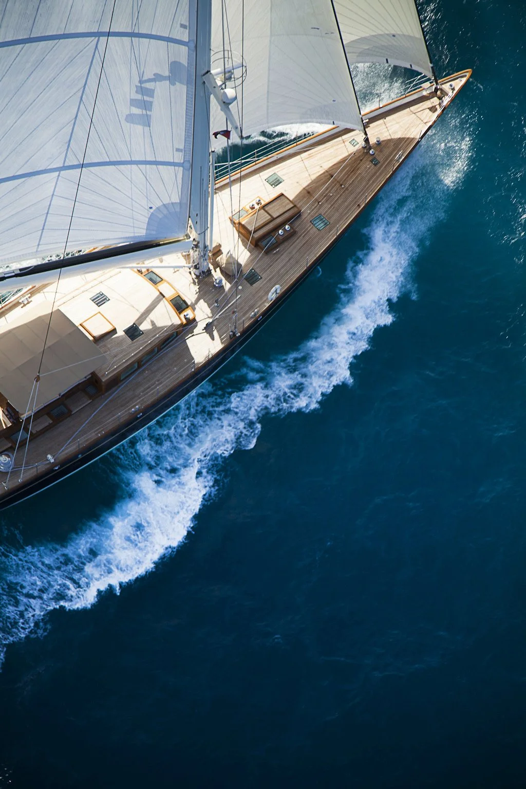 Overhead view of a sailing yacht with white sails and a wooden deck, cutting through blue ocean water, leaving a wake behind.