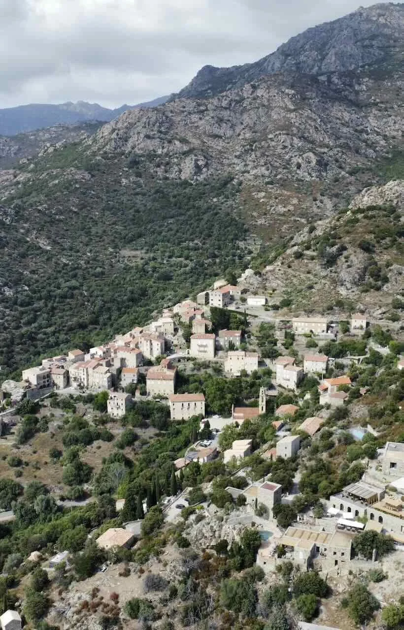 A hillside village with stone buildings, terracotta roofs, and winding roads surrounded by green vegetation and rocky mountains under a cloudy sky.