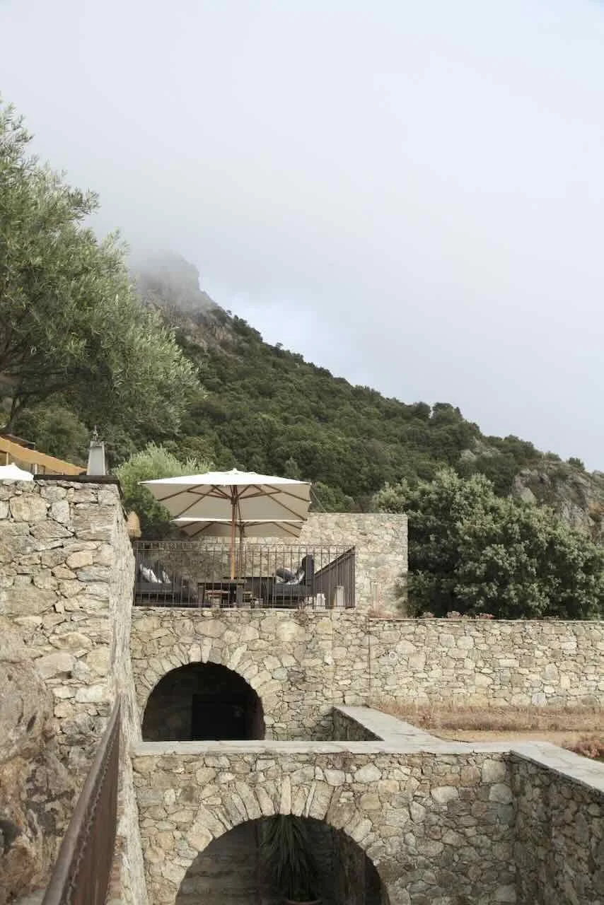 Stone terrace with outdoor seating and a large white patio umbrella, overlooking a mountain landscape with trees and fog.