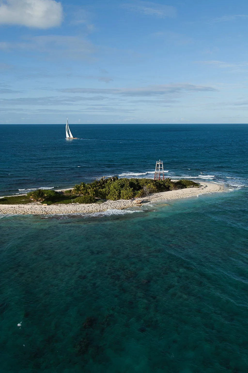 A small island with greenery and a lifeguard tower, surrounded by turquoise waters. A sailboat is visible in the distance on the ocean under a blue sky with scattered clouds.