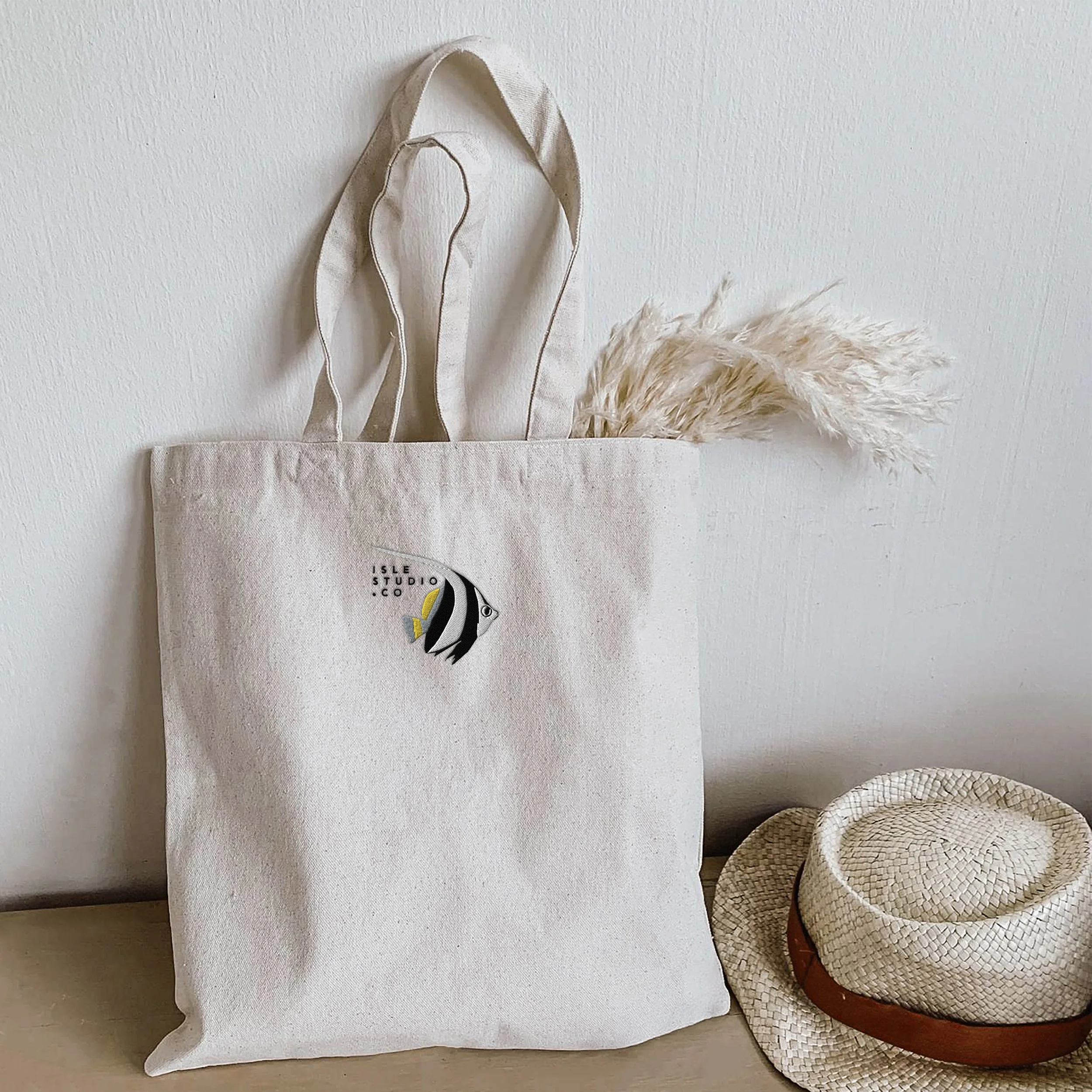 Cream-colored tote bag with a small fish logo and text 'ISLE STUDIO.CO' embroidered on it, placed against a white wall, with a beige straw hat with a brown band resting on the surface next to it, and dried pampas grass in the tote bag.