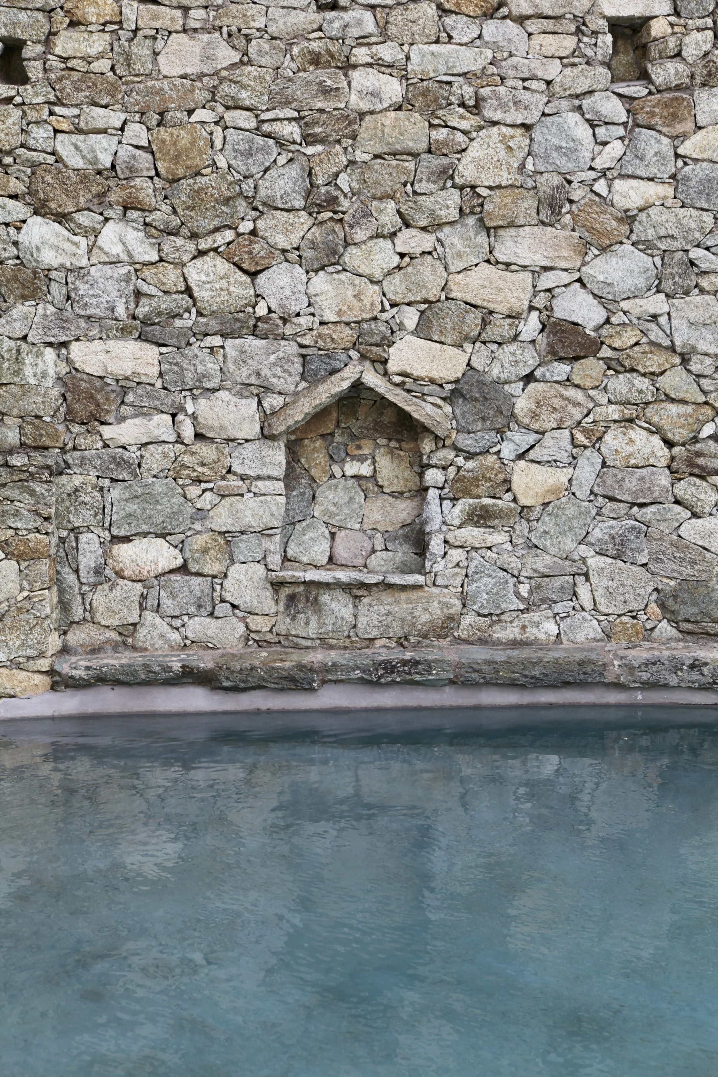 A stone wall with a small, house-shaped niche and a ledge above a body of water.