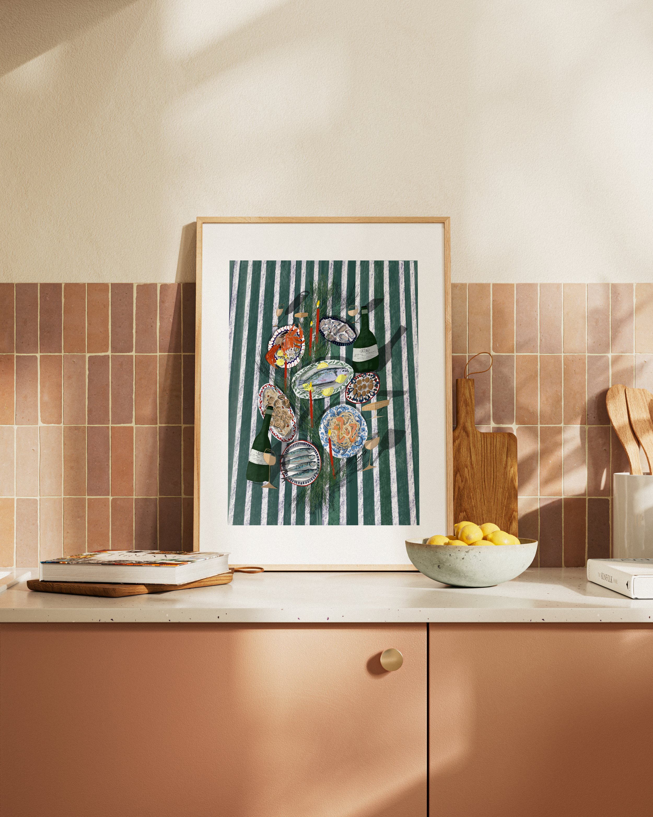 Kitchen countertop with a framed artwork of a table setting, a bowl of lemons, a cutting board, and books, against a tiled wall with shadow lighting.