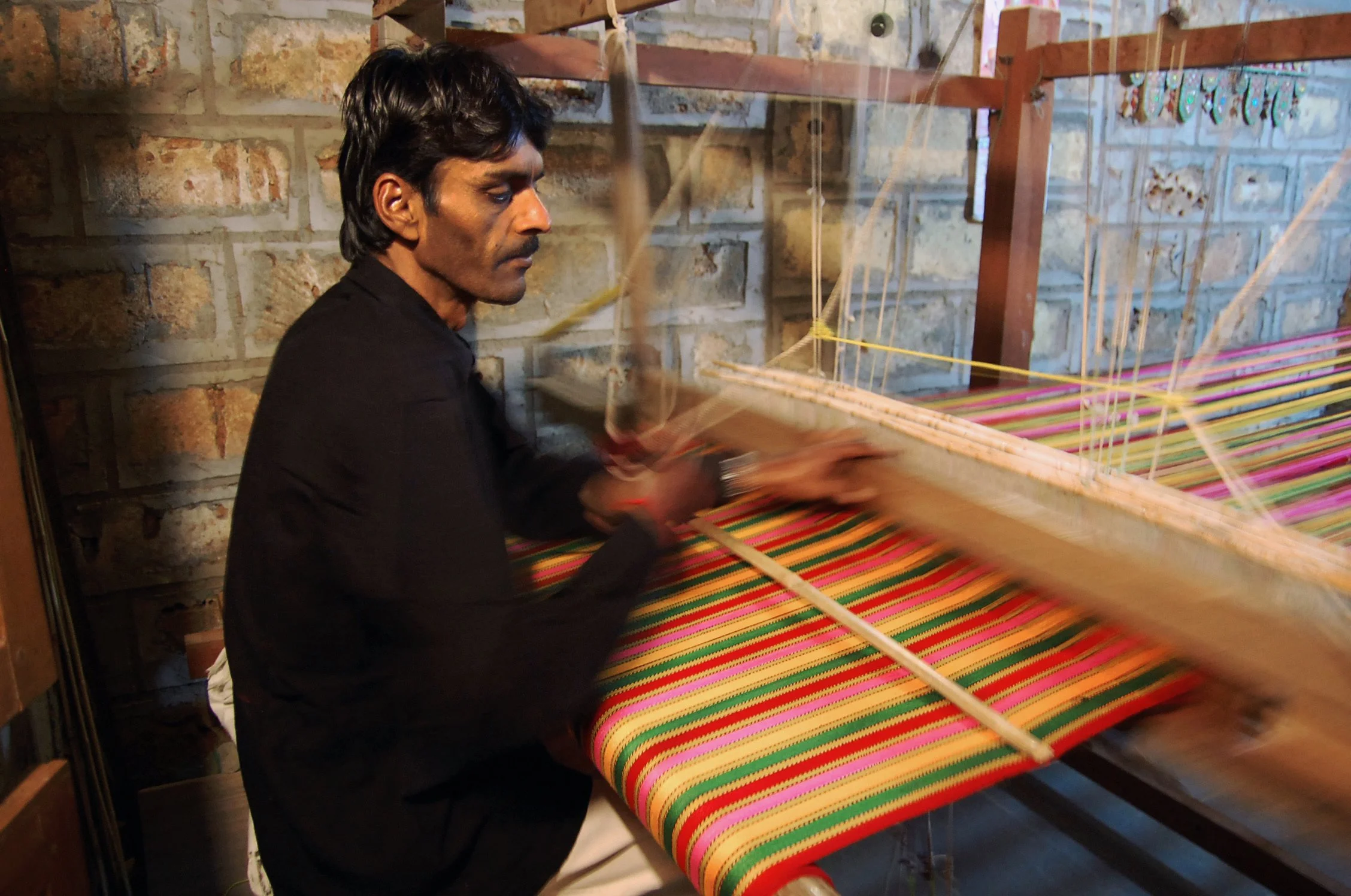 A man working on a traditional handloom with colorful striped fabric in a rustic room with brick walls.