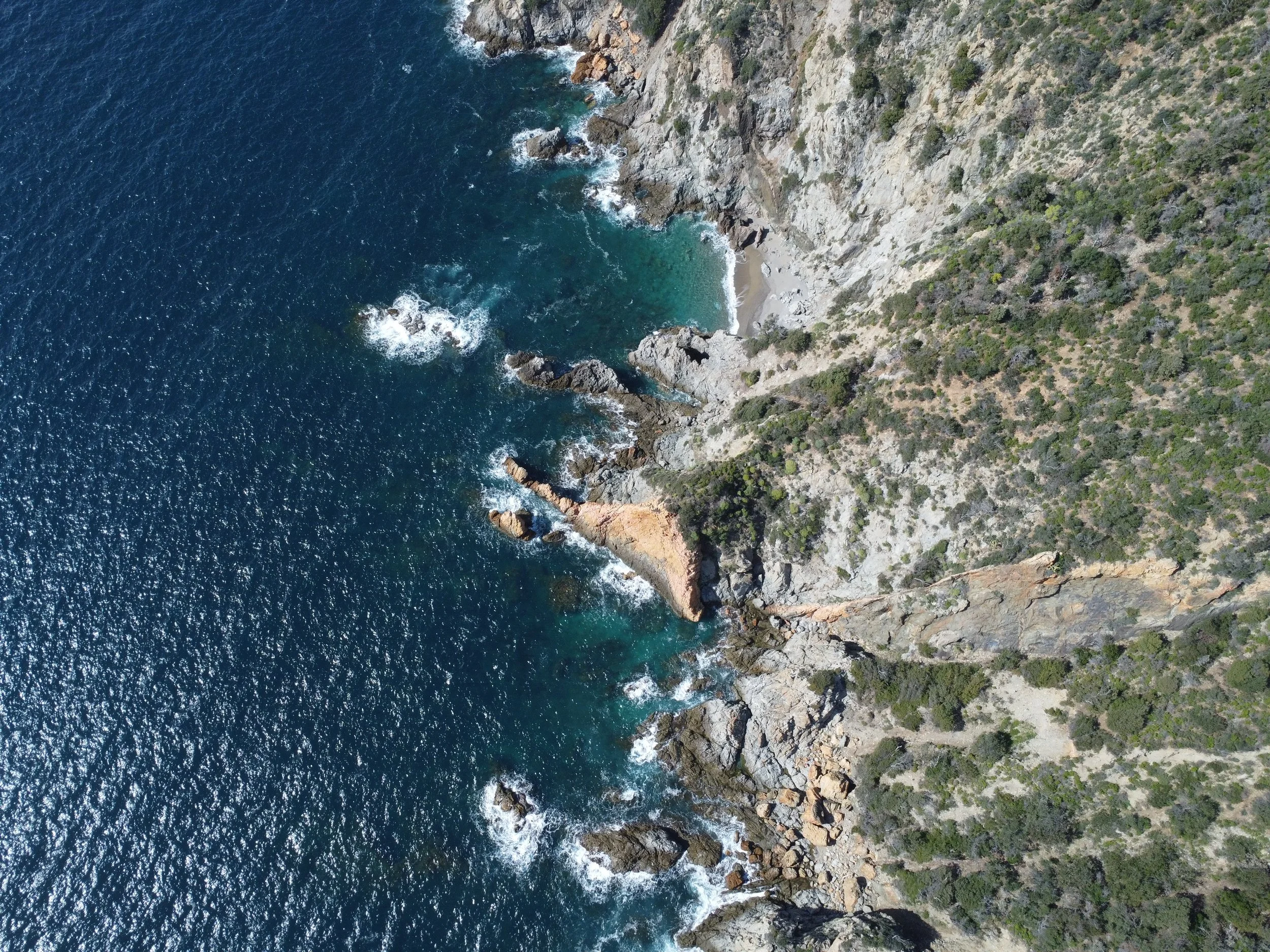 Aerial view of a rugged coastline with a narrow beach, rocky cliffs, and green shrubbery meeting the deep blue ocean.