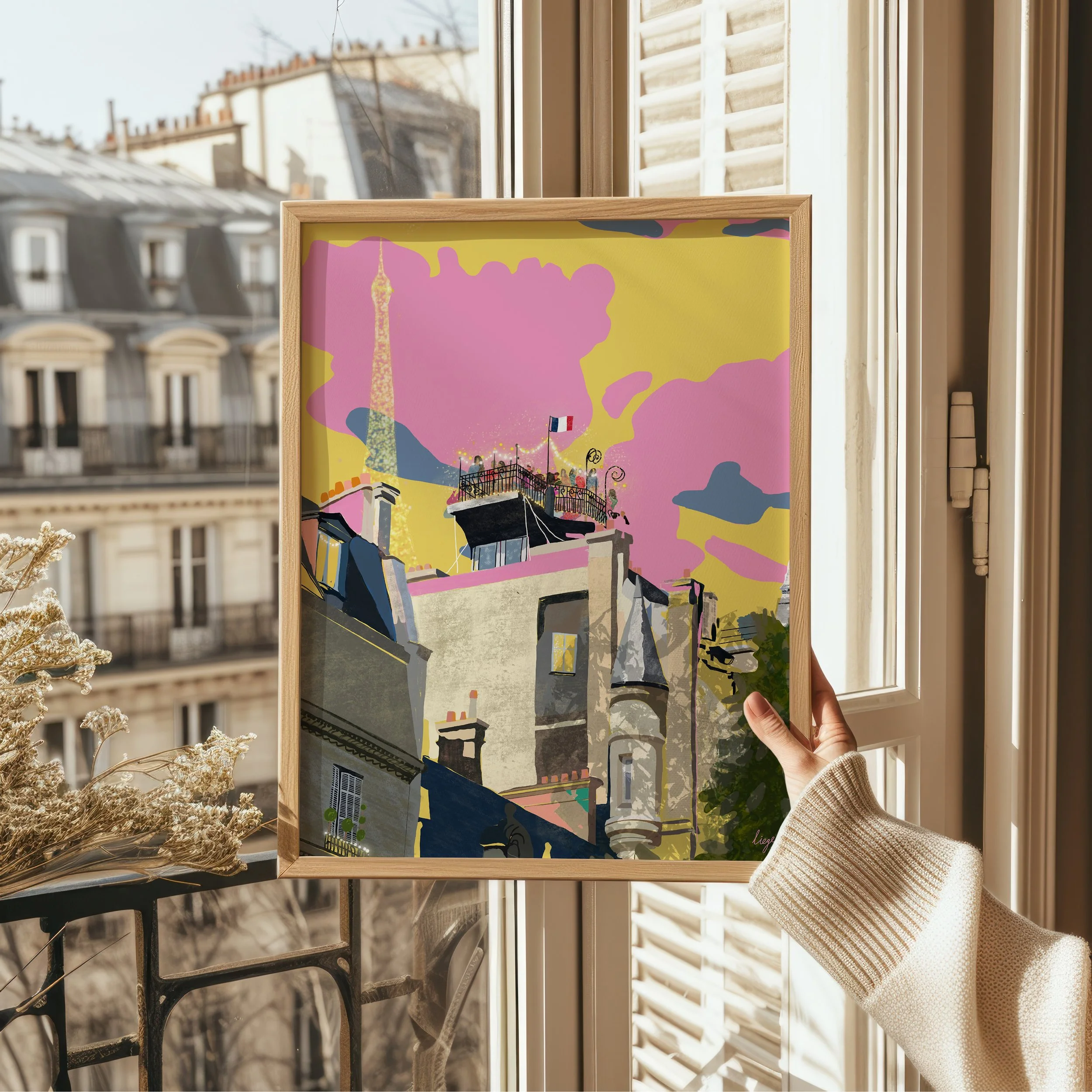 A person holding a framed painting of Paris rooftops with the Eiffel Tower in the background, near a window with sunlight.