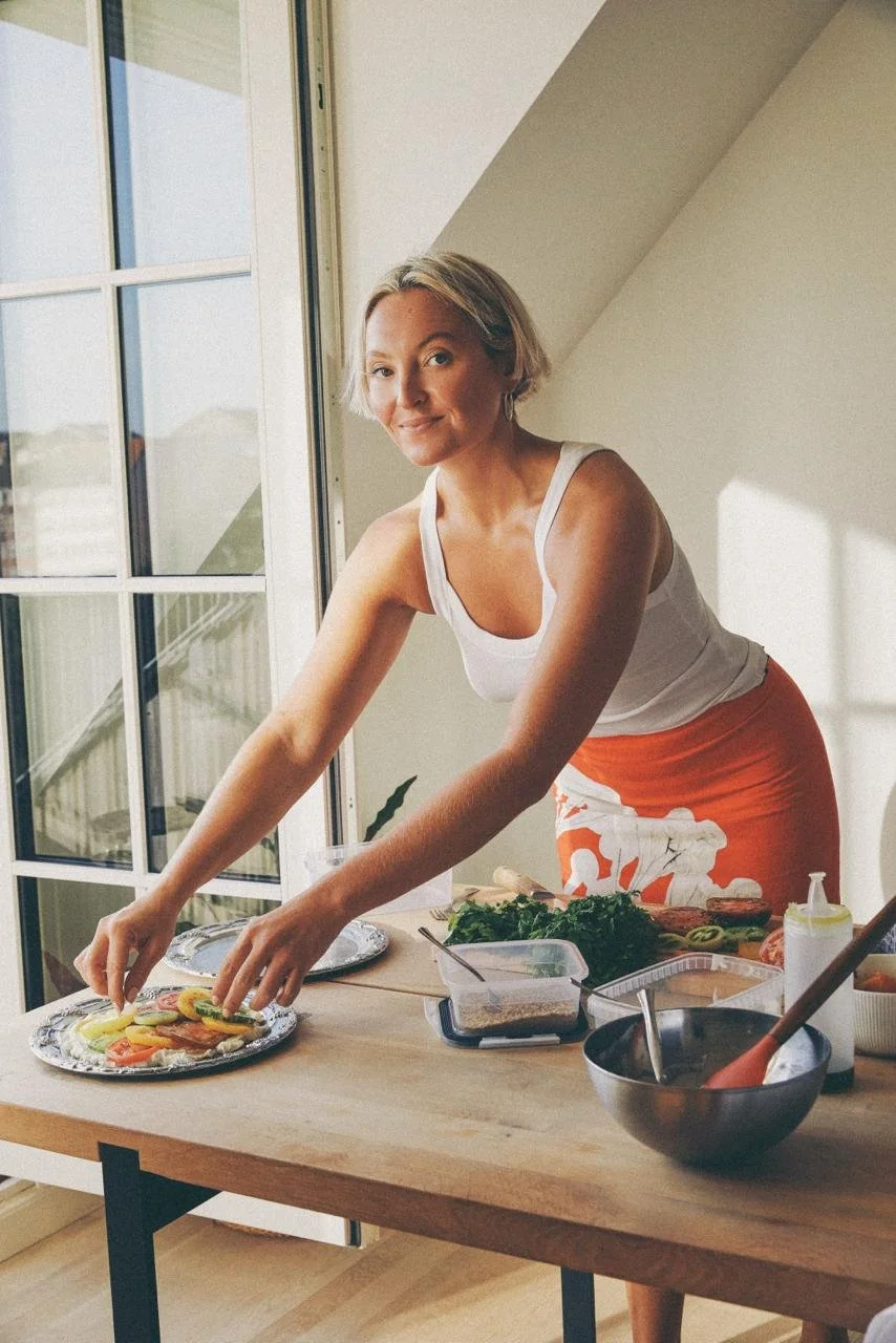 A woman preparing a colorful vegetable platter or salad on a table near a large window in a bright room.
