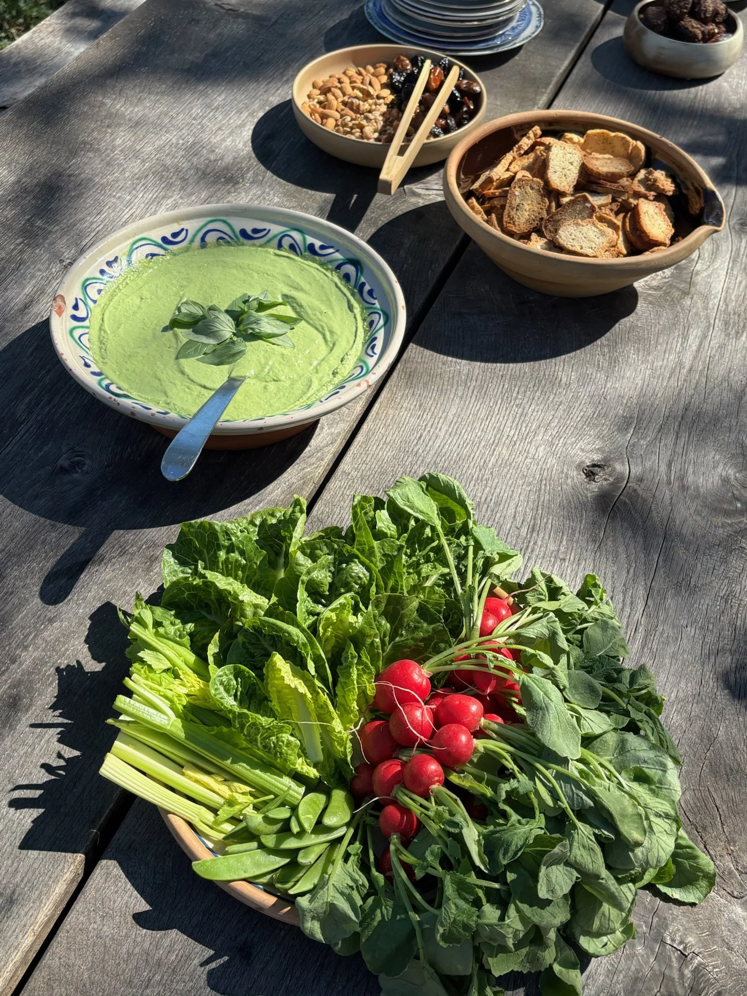 Multiple bowls and plates of salad and cooked vegetables on a light-colored table, with a person serving food in the background near a window.