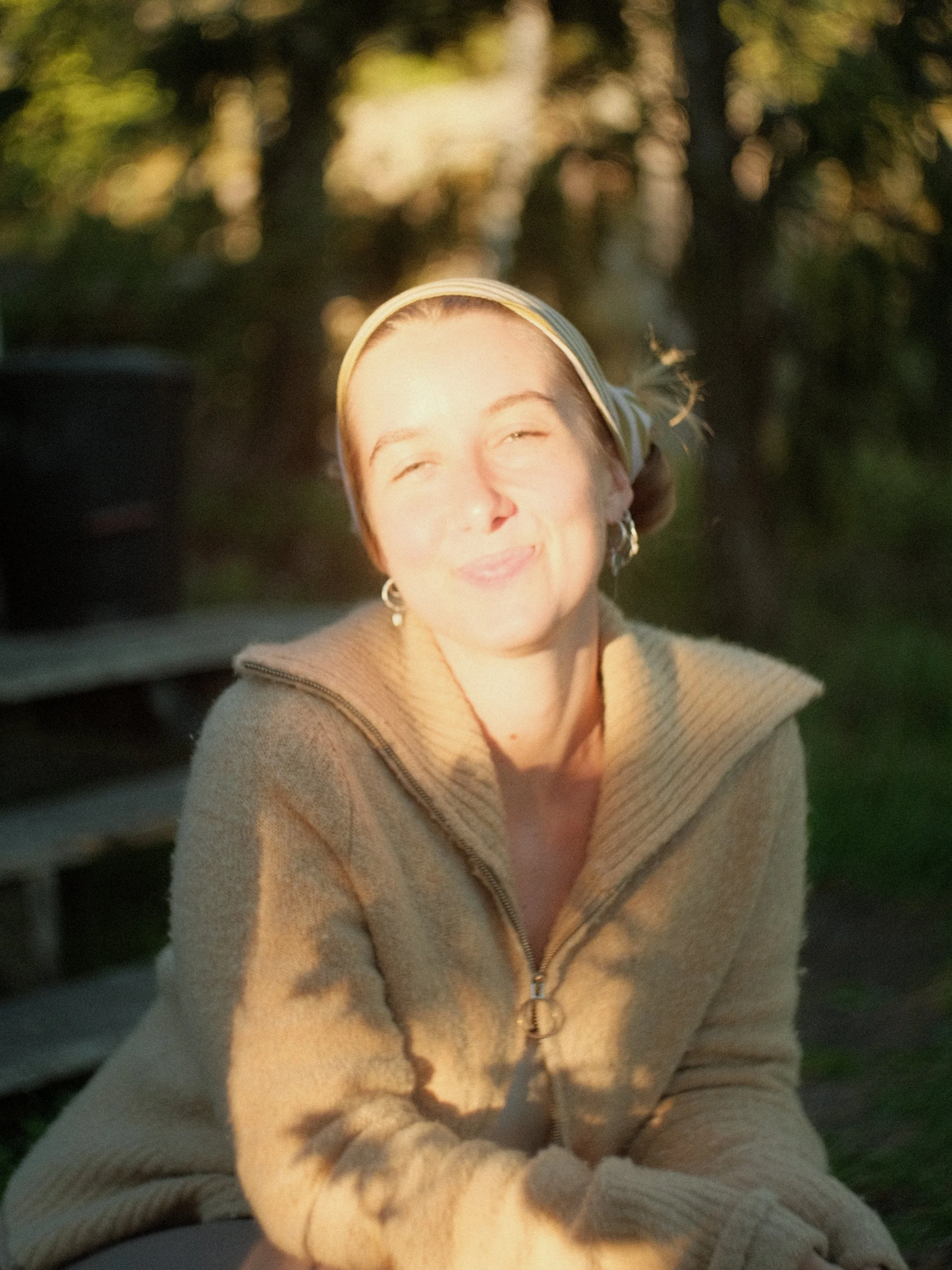 A woman with light skin, brown hair, and hoop earrings is smiling with her eyes almost closed. She is wearing a beige zip-up jacket and a yellow and white striped headband. The background features blurred trees and sunlight.