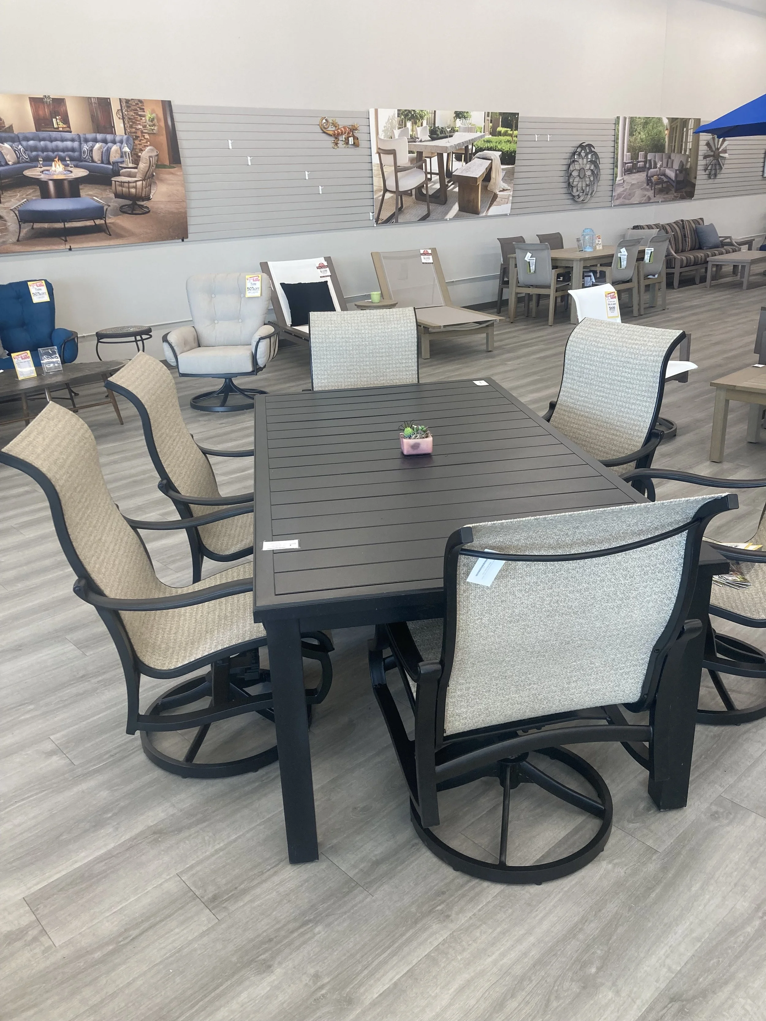 A furniture showroom displays various chairs, tables, and outdoor furniture, with a black dining table and multiple beige and patterned chairs in the foreground.