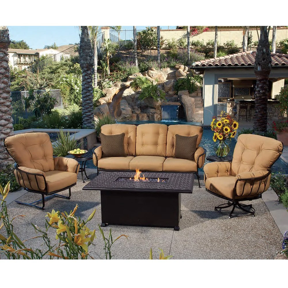 Outdoor patio set with tan cushioned chairs, sofa, and a fire pit table in a garden setting with palm trees, water feature, and sunflowers.