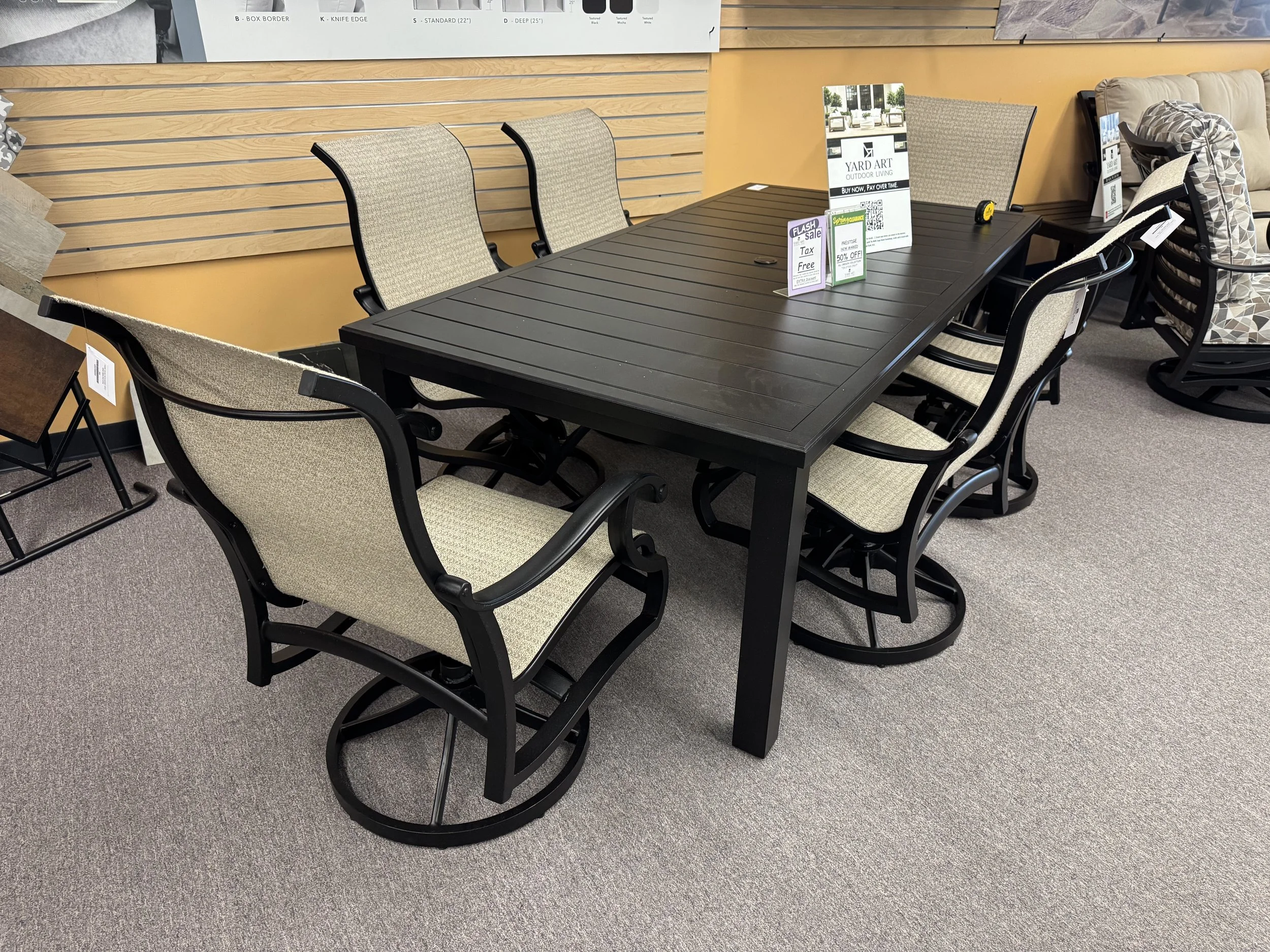A black outdoor dining table with six light-colored cushioned chairs in a furniture store.