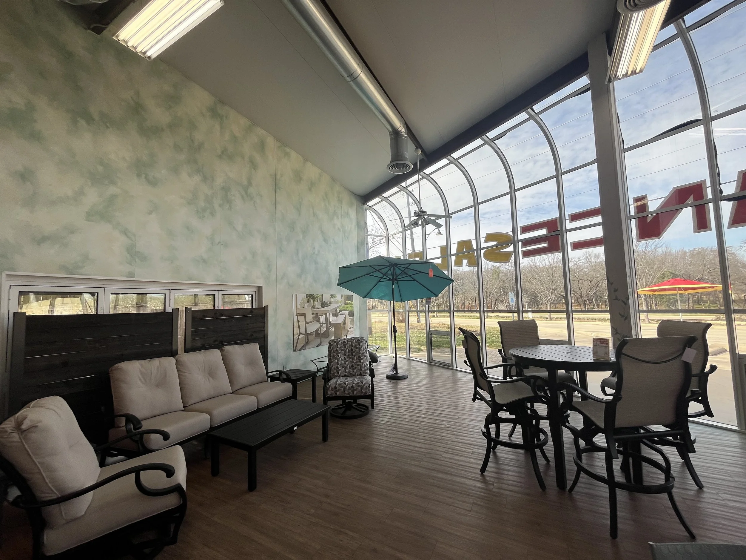 Interior of a cafe with beige cushioned seating, a round table with chairs, a blue patio umbrella, large windows, and a view of outdoors with trees and a yellow umbrella.