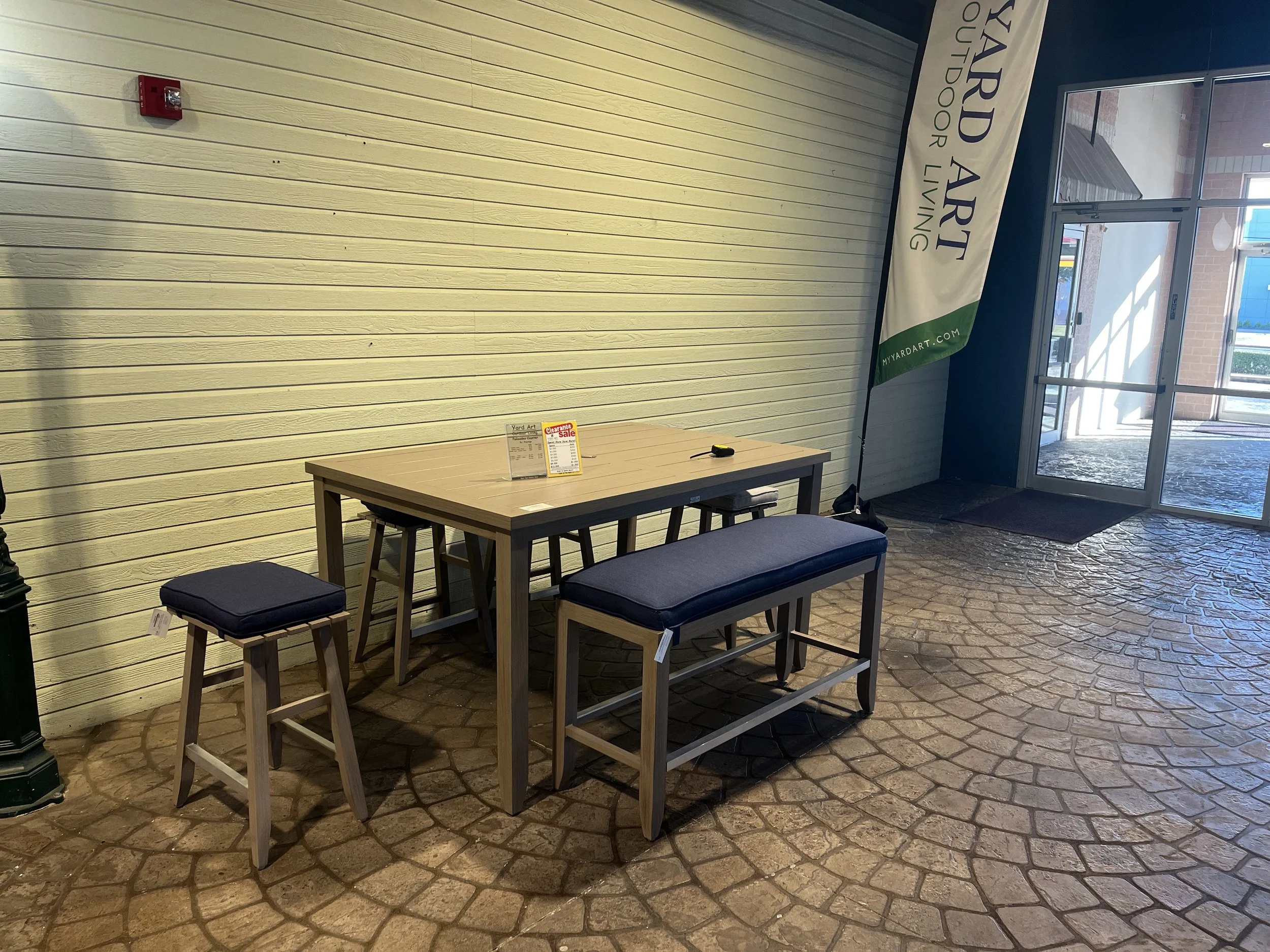 Indoor seating area with a rectangular table, three bar stools with dark cushions, a bench with a dark cushion, and a banner that reads "Yard Art" near an entrance door with glass panels. The floor has a stone pattern and the wall is painted light yellow.