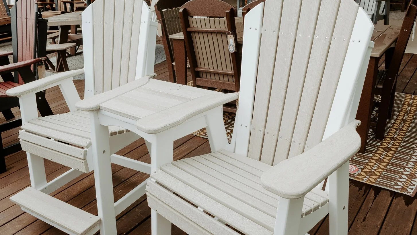 Two white Adirondack rocking chairs with built-in table on wooden deck, surrounded by other outdoor furniture.