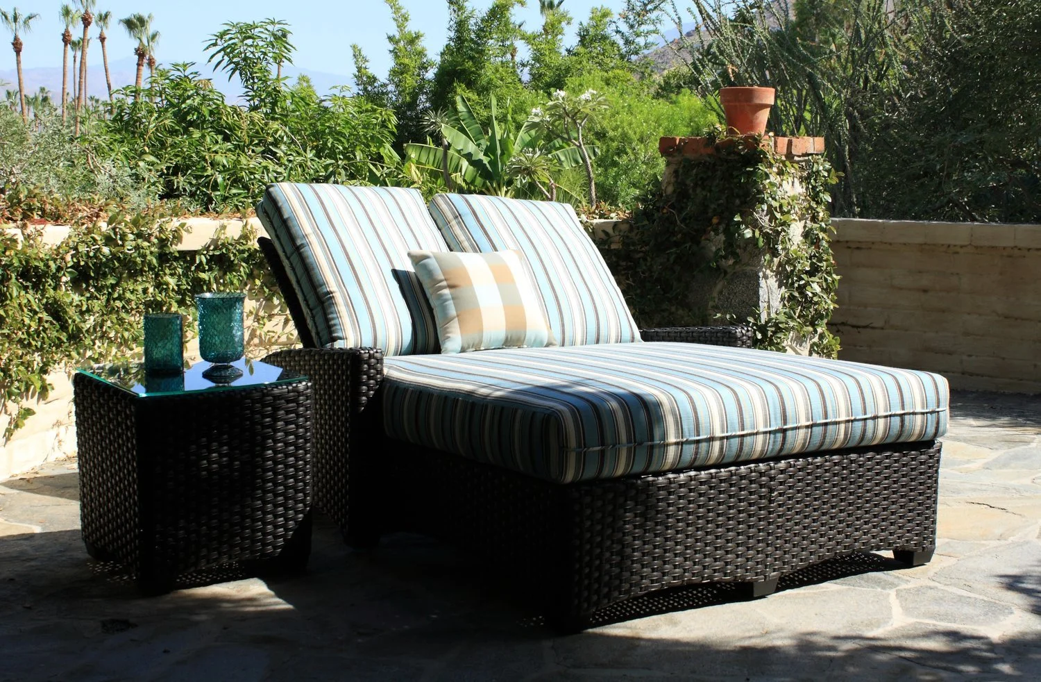 Outdoor patio with wicker lounge chair and striped cushions next to a small table with turquoise candles, surrounded by greenery.