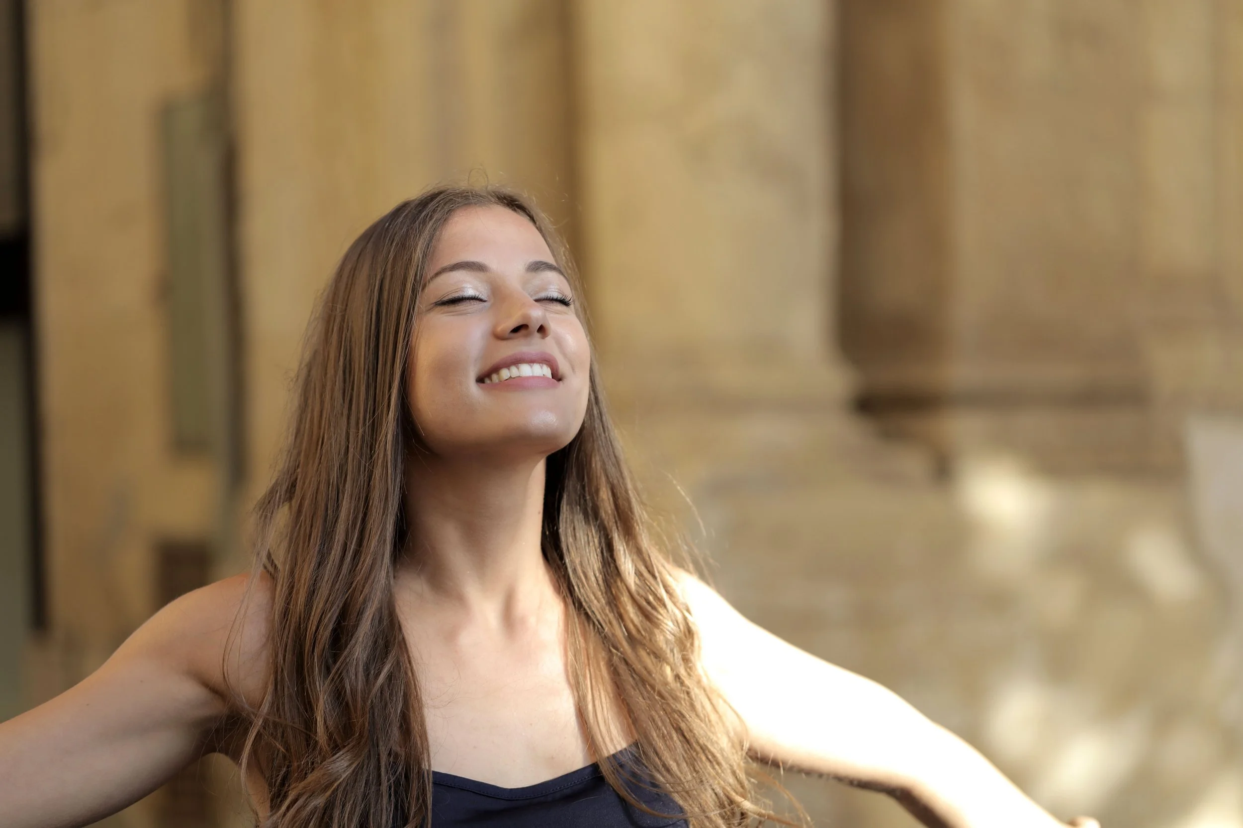 A woman with long brown hair smiling with her eyes closed in front of a blurred background.