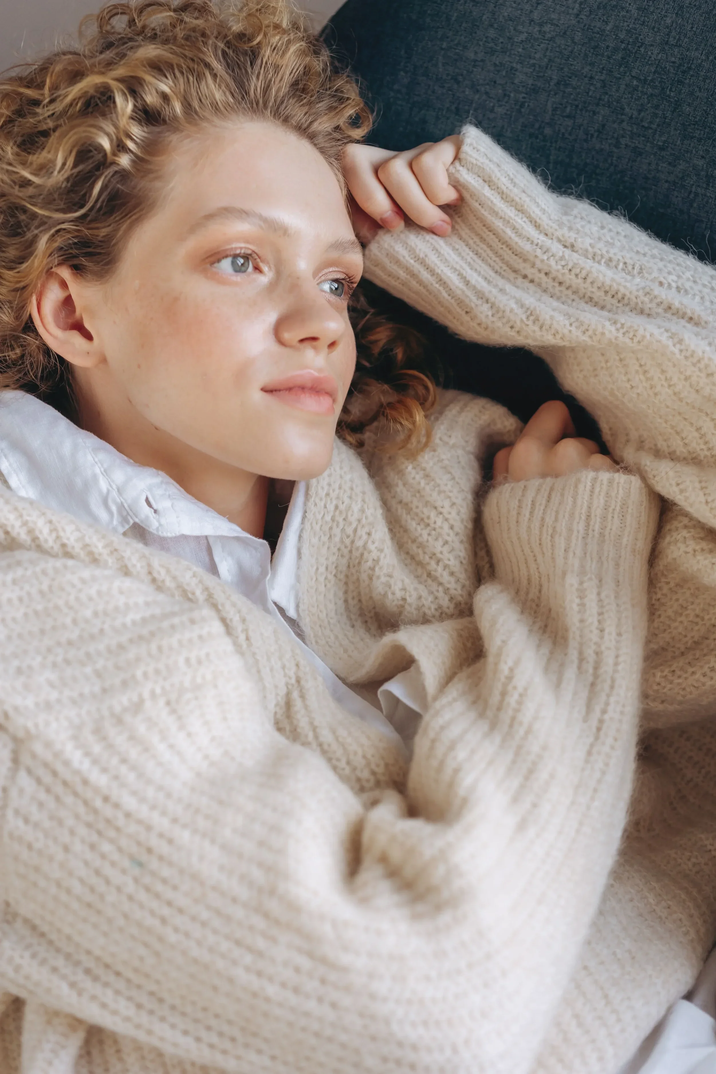 Close-up of a young woman with curly red hair, lying on her side on a dark grey couch, wearing a white shirt and a cream-colored knit sweater. She appears relaxed with a peaceful expression.