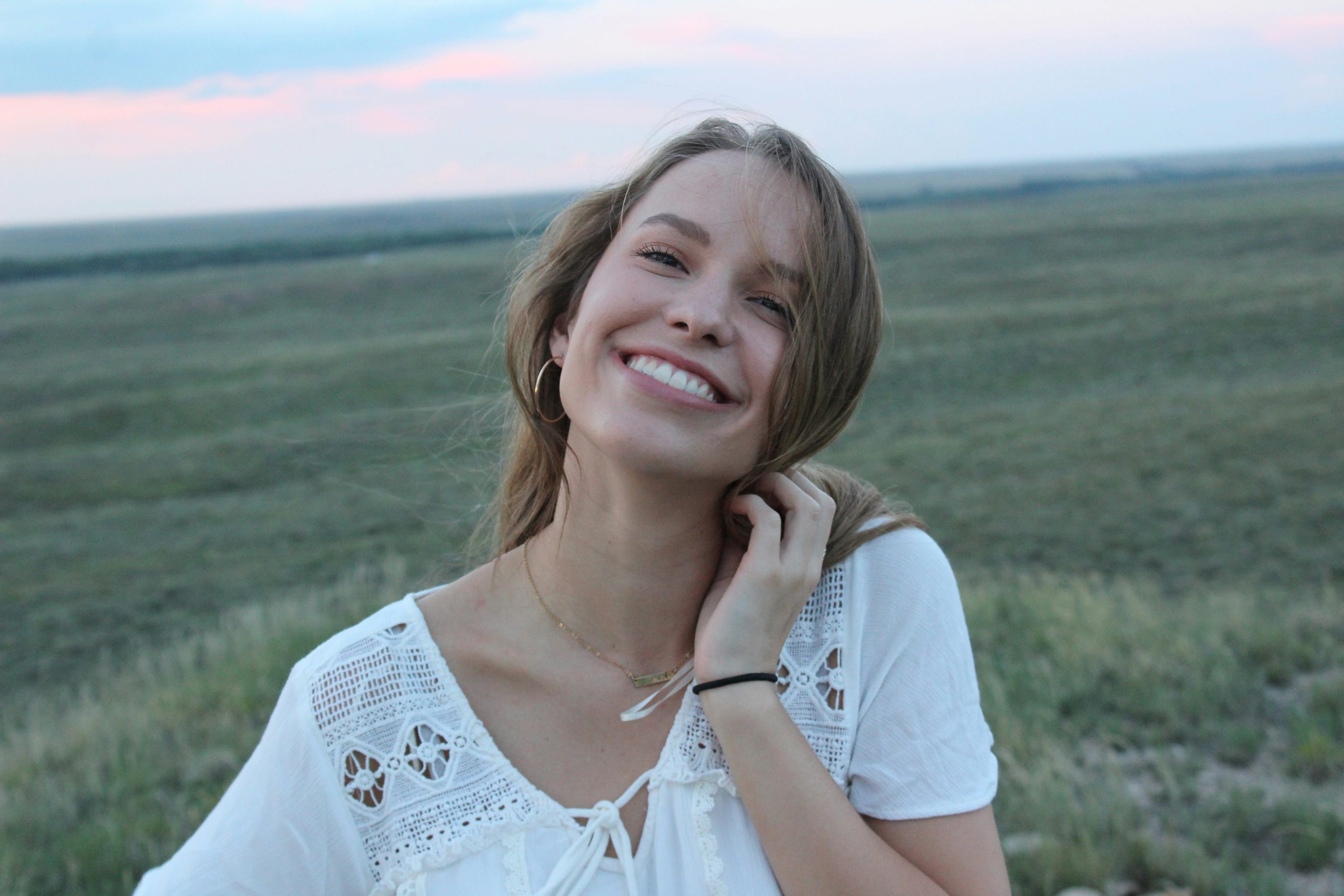 A young woman with light brown hair, smiling and wearing hoop earrings and a white lace blouse, standing outdoors in a green field under a sky with pink and blue clouds.