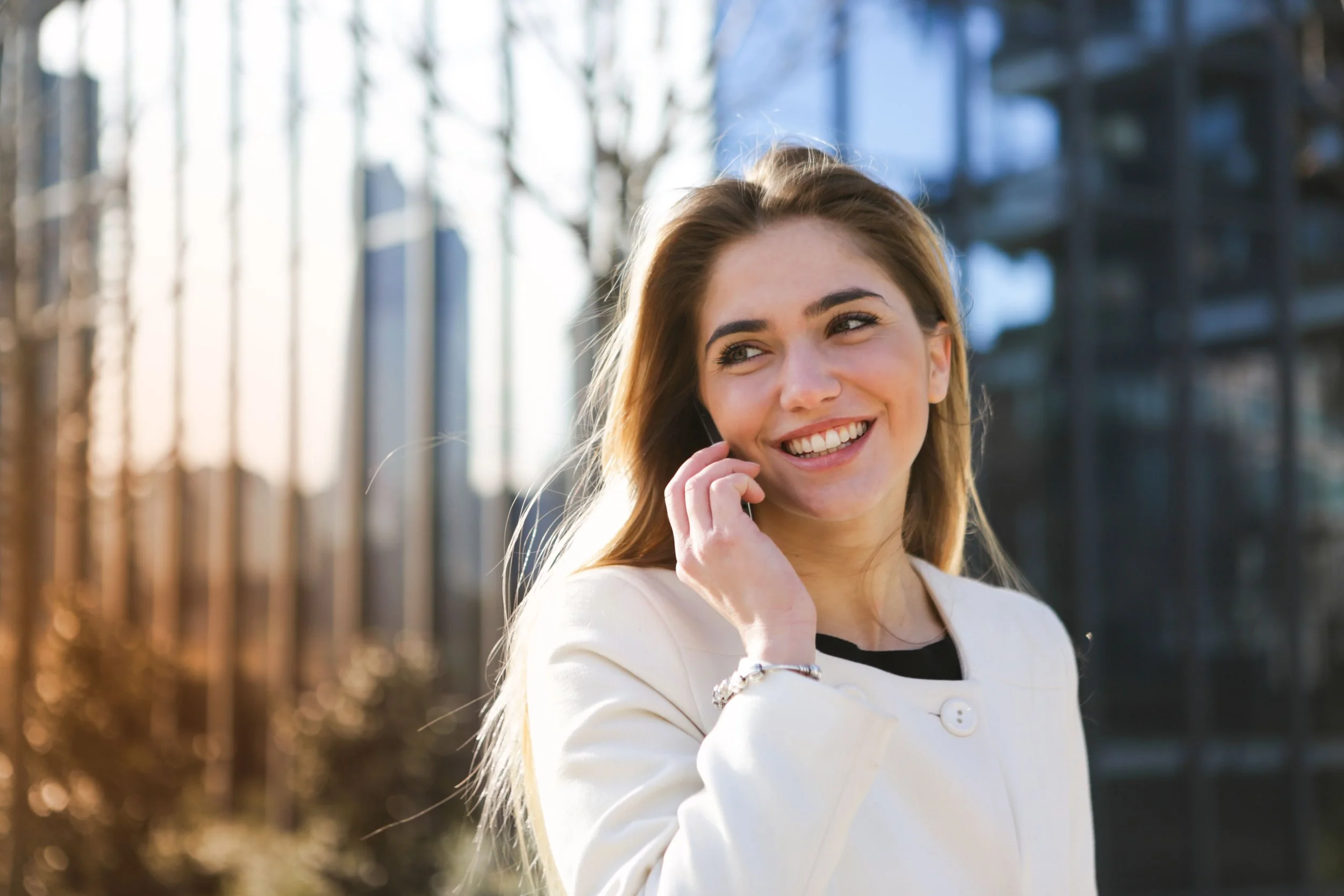 A smiling young woman with long hair talking on her mobile phone outdoors on a sunny day, with a blurred background of trees and buildings.