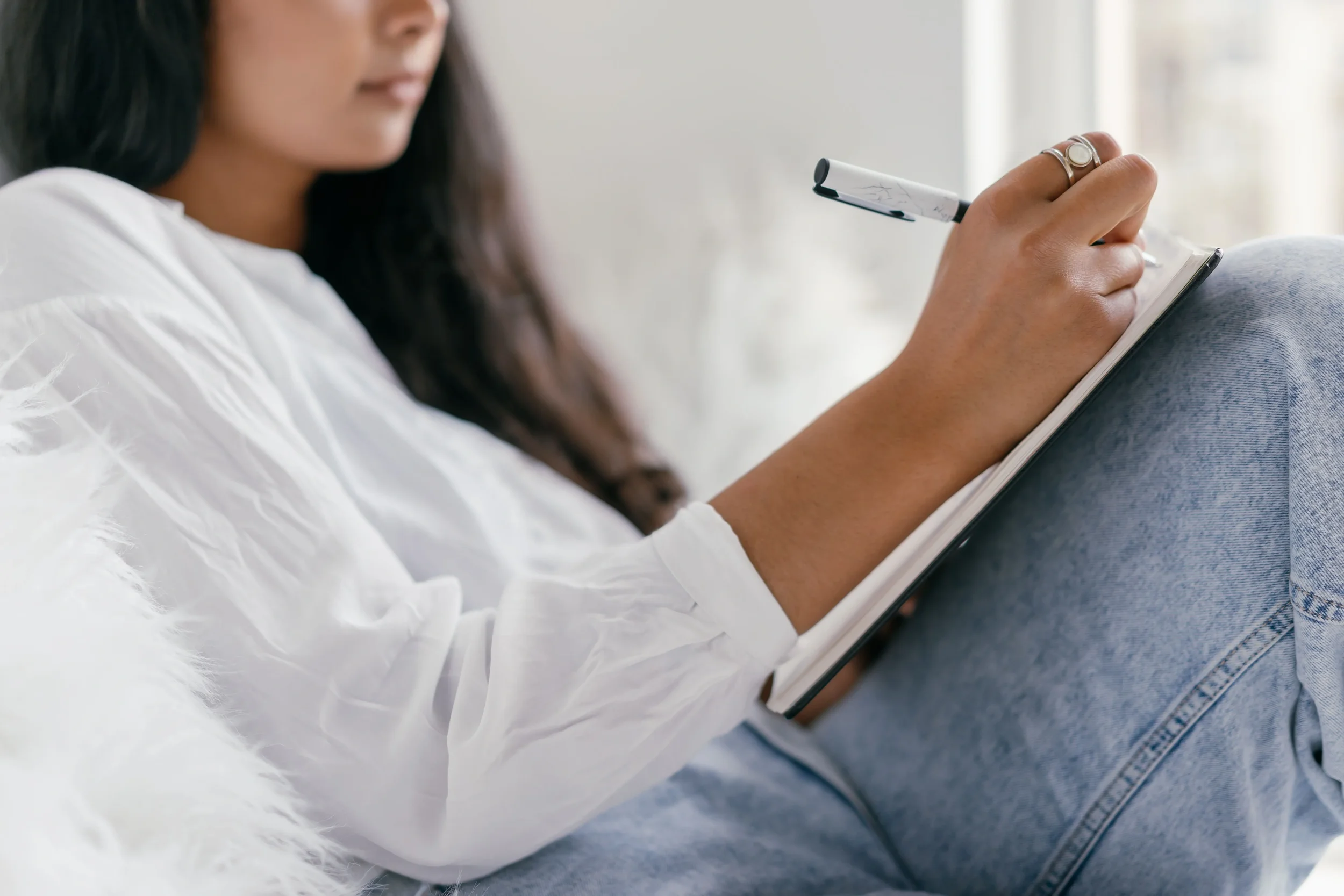 Close-up of a woman with dark hair writing in a notebook with a white marker while sitting in casual clothes.