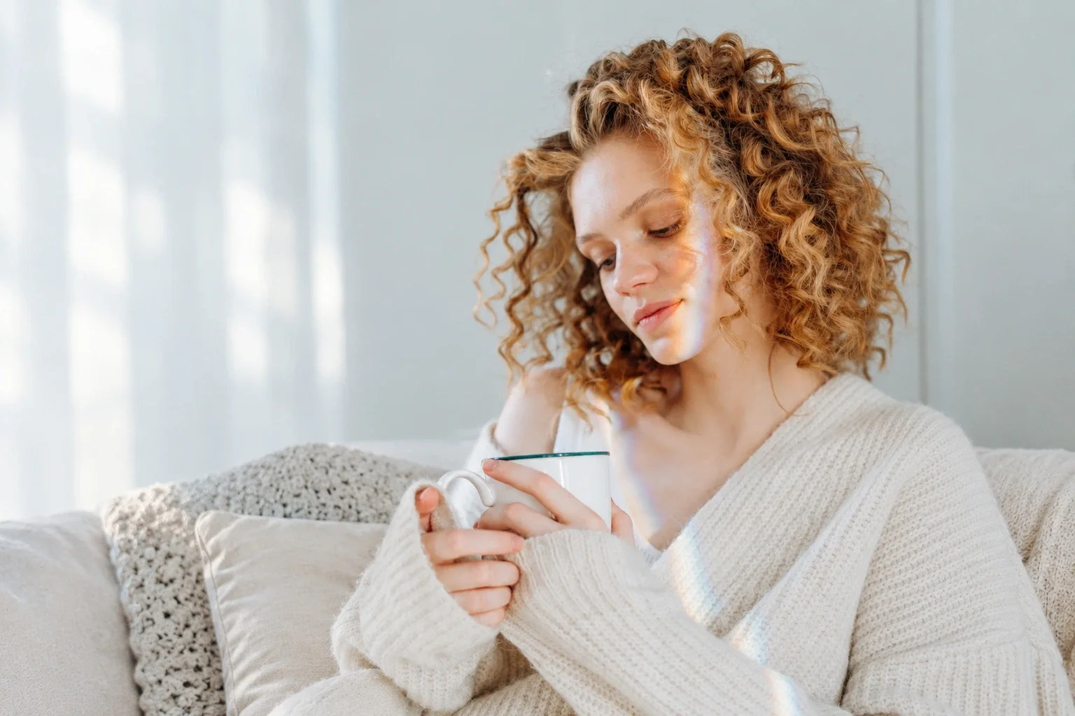 A young woman sitting on a sofa, holding a mug, wearing a cream knit sweater, with sunlight streaming in through a window.