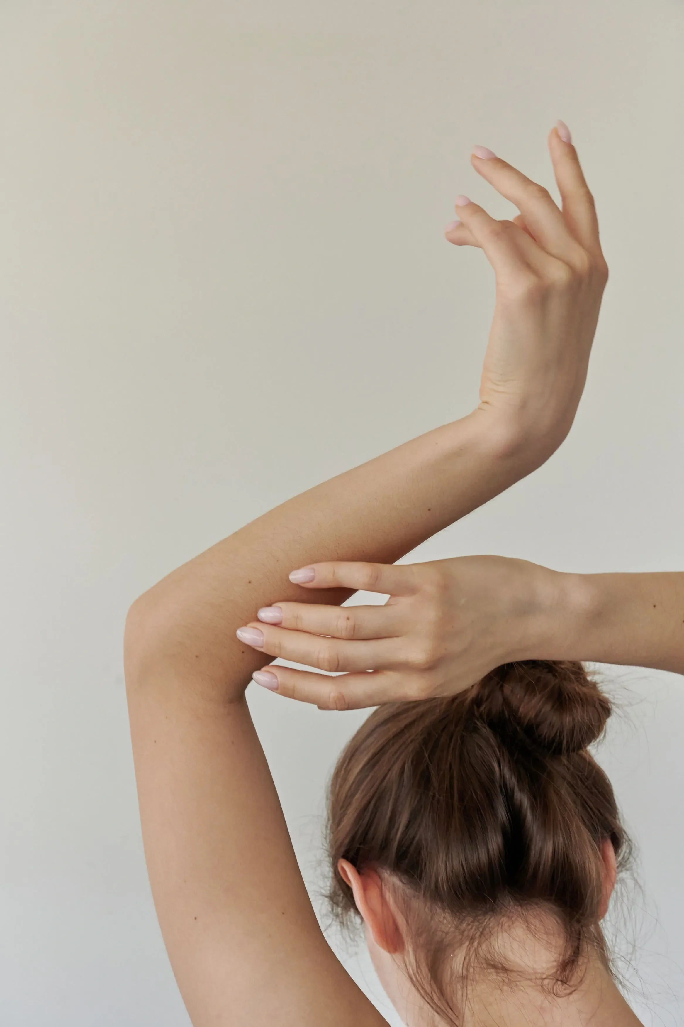 A person with brown hair styled in a bun is posing with their arm bent and hand gracefully extended, against a plain light-colored wall background.