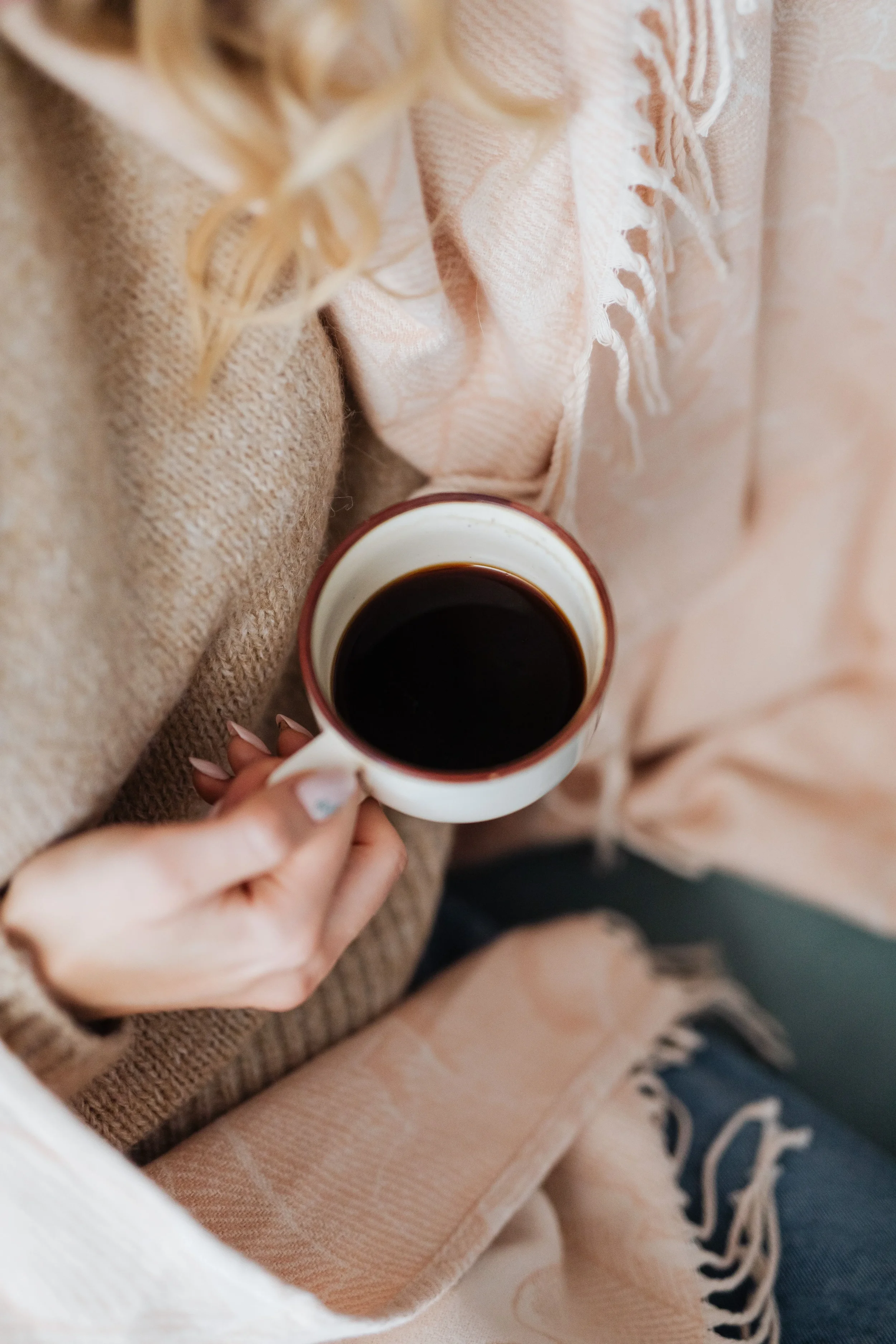 A person holding a mug of black coffee, resting on their lap, wearing a beige sweater and a patterned blanket.