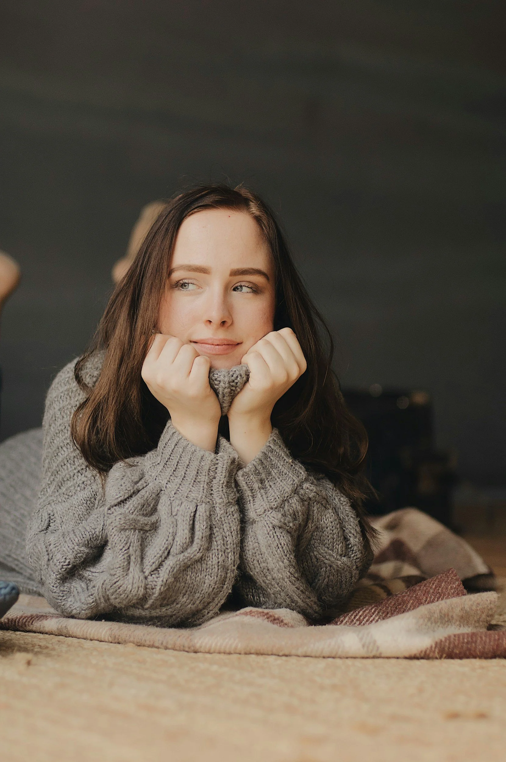 A young woman lies on her stomach on a bed, resting her chin on her hands, looking thoughtfully to the side. She has long brown hair and is wearing a cozy gray knit sweater.
