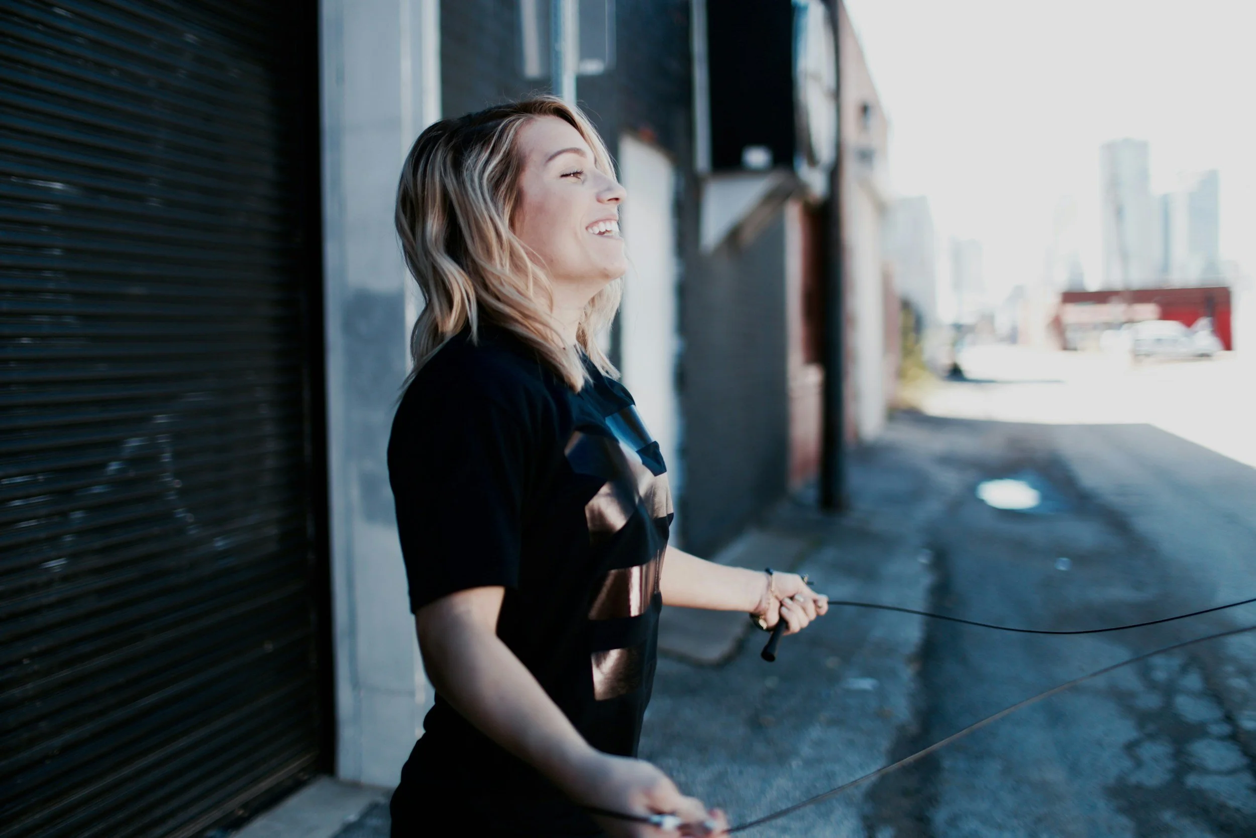 A woman with blonde hair smiling and jumping rope on an urban sidewalk.