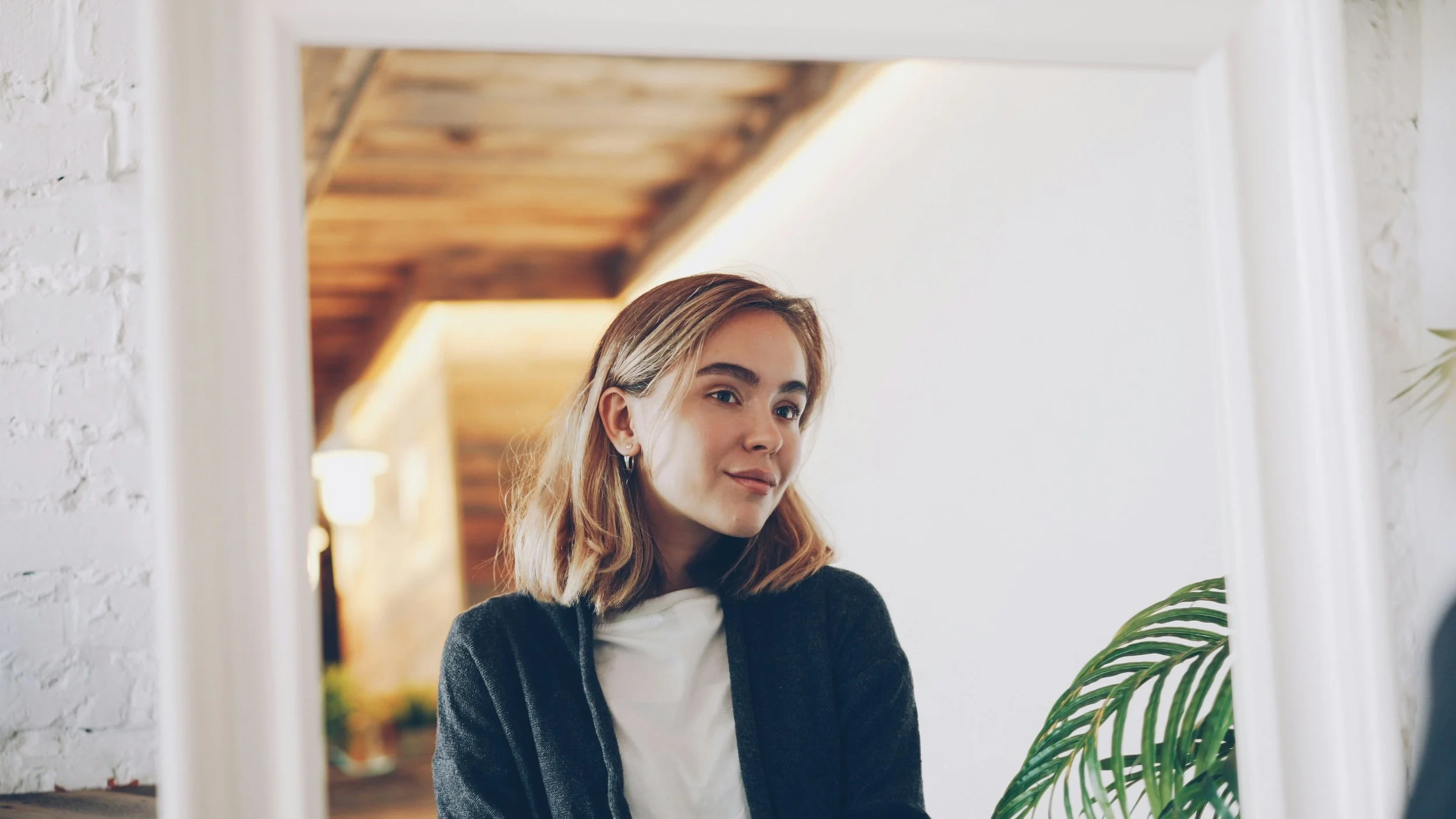 A young woman with shoulder-length blonde hair looking at herself in a mirror with a white frame, in a well-lit room with a brick wall and wooden ceiling in the background.