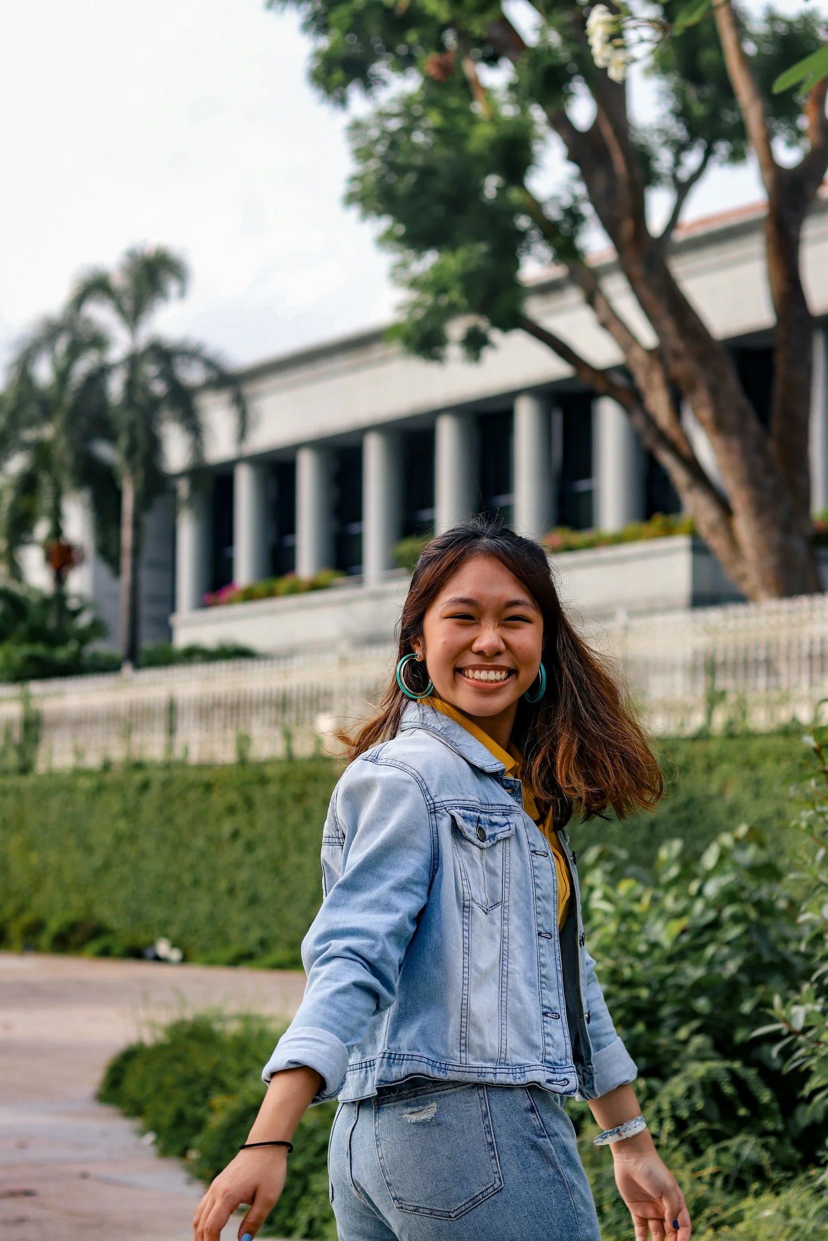 A young woman with long brown hair smiling and walking outdoors in front of a building with tall columns, a tree, and greenery.