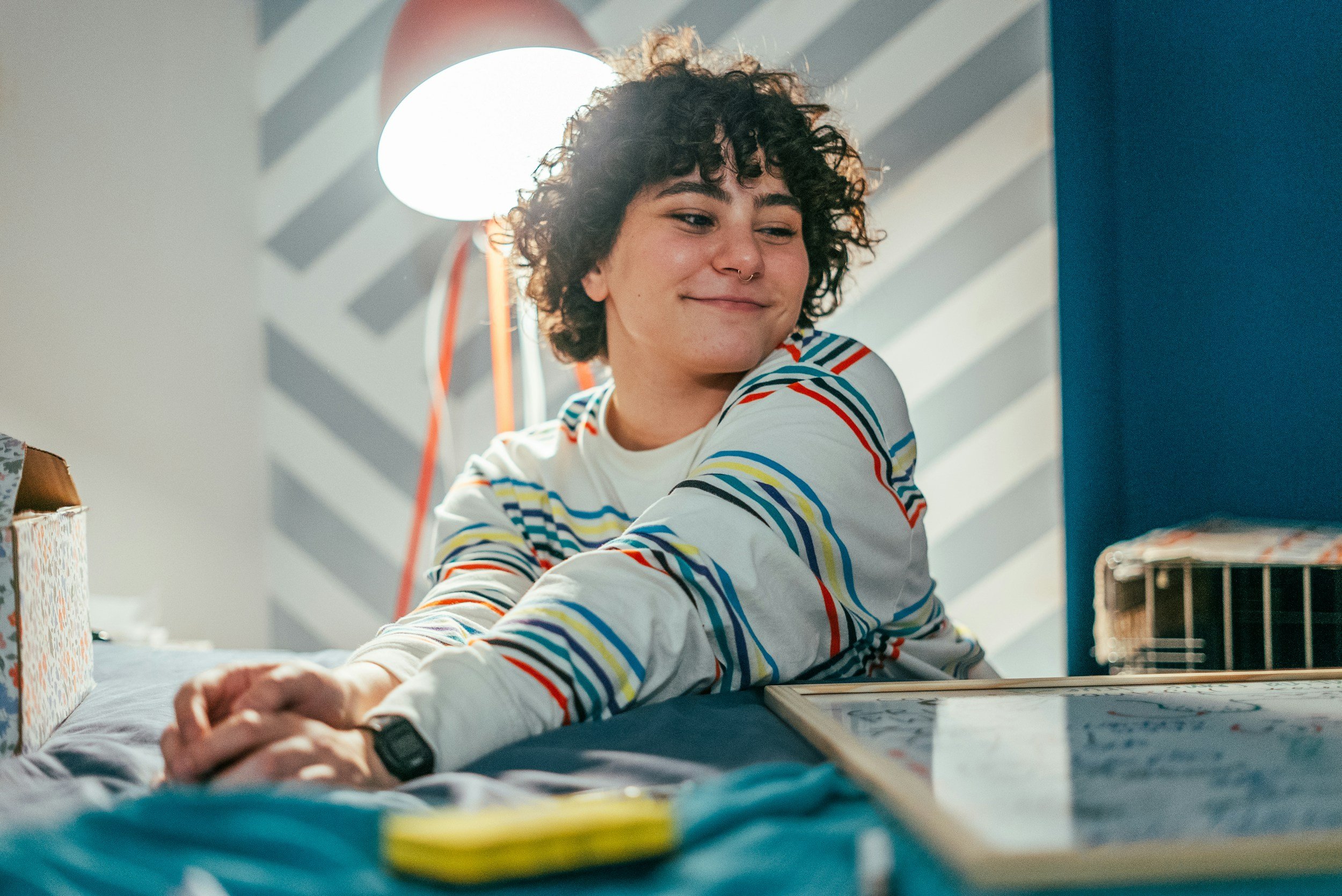 A young person with curly hair wearing a striped long-sleeve shirt, smiling and looking to the side, sitting at a table in a room with a blue and gray wall, a bright light in the background, and various objects on the table.